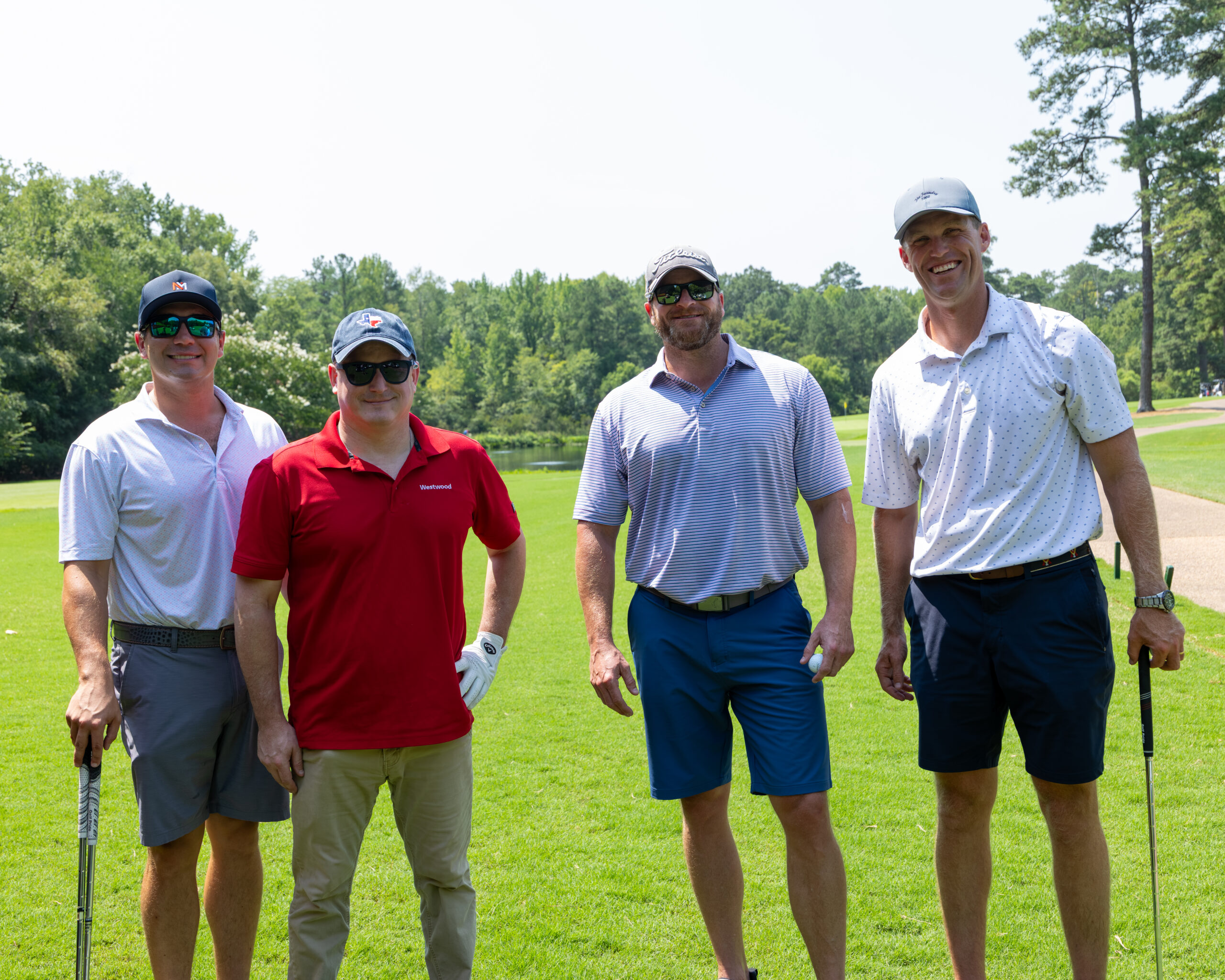 Four men in golf attire stand on a grassy field near trees.