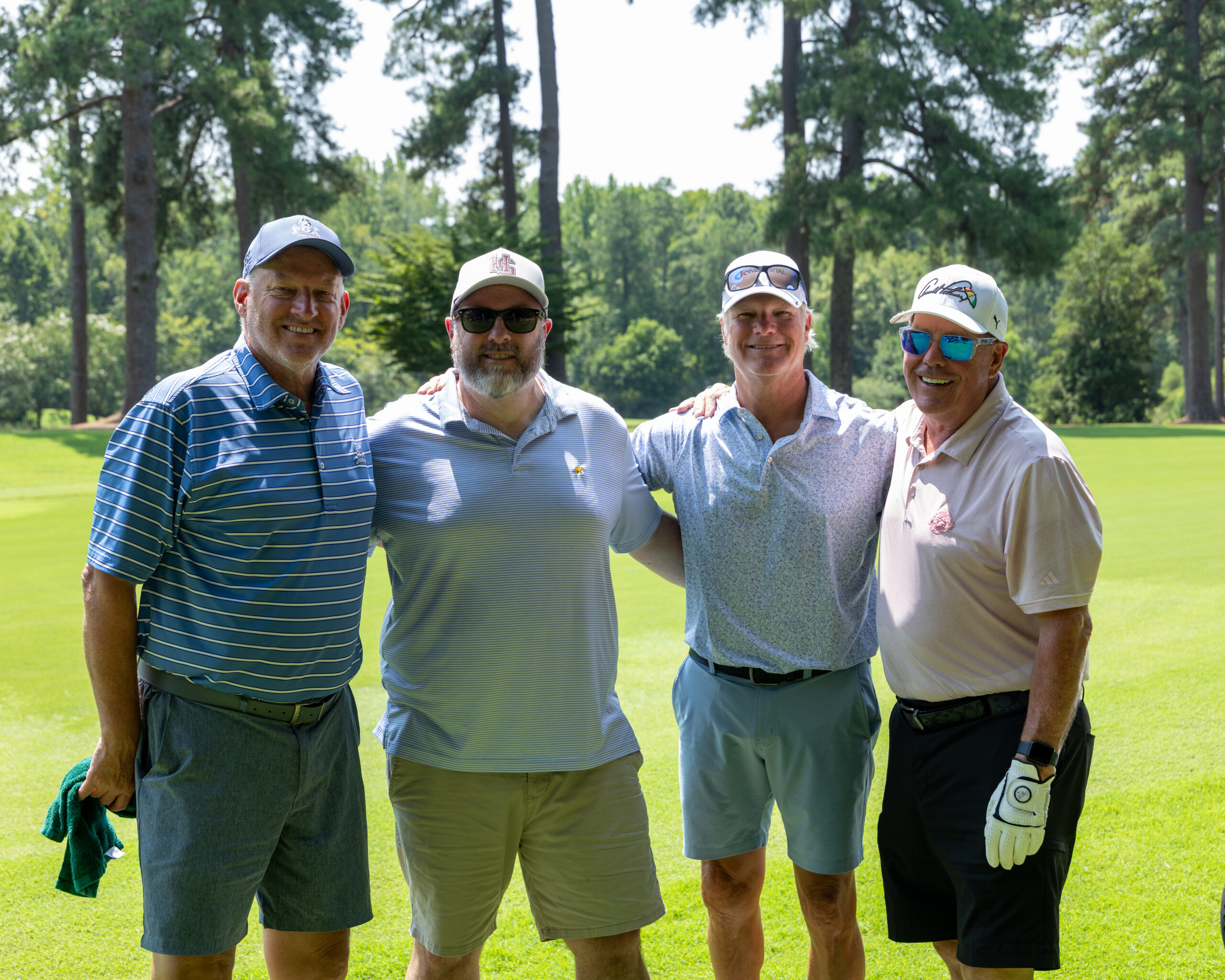 Four men in golf attire standing together and smiling outdoors on a grassy field near trees.