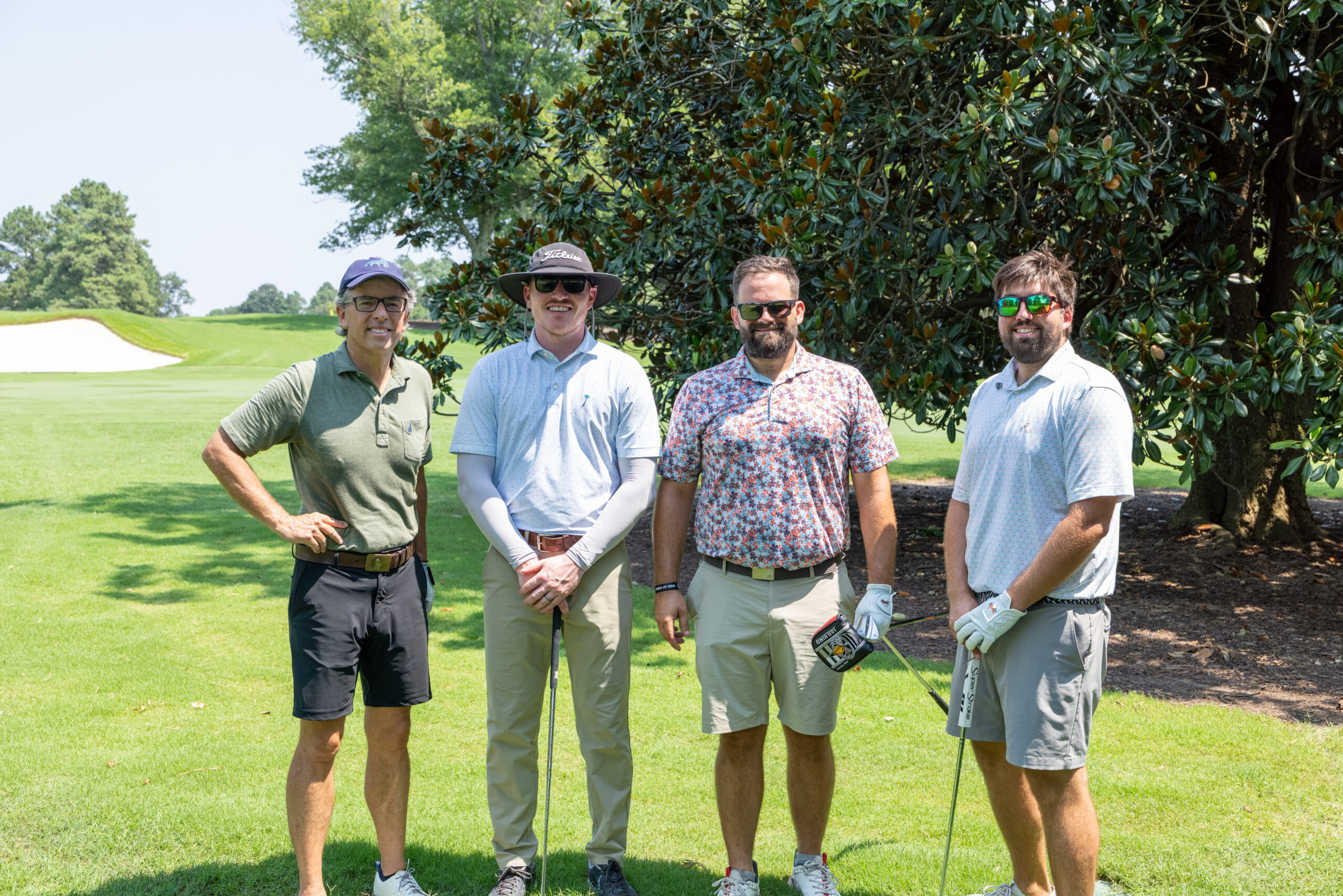 Four men in golf attire standing together on a lush green golf course next to large trees.