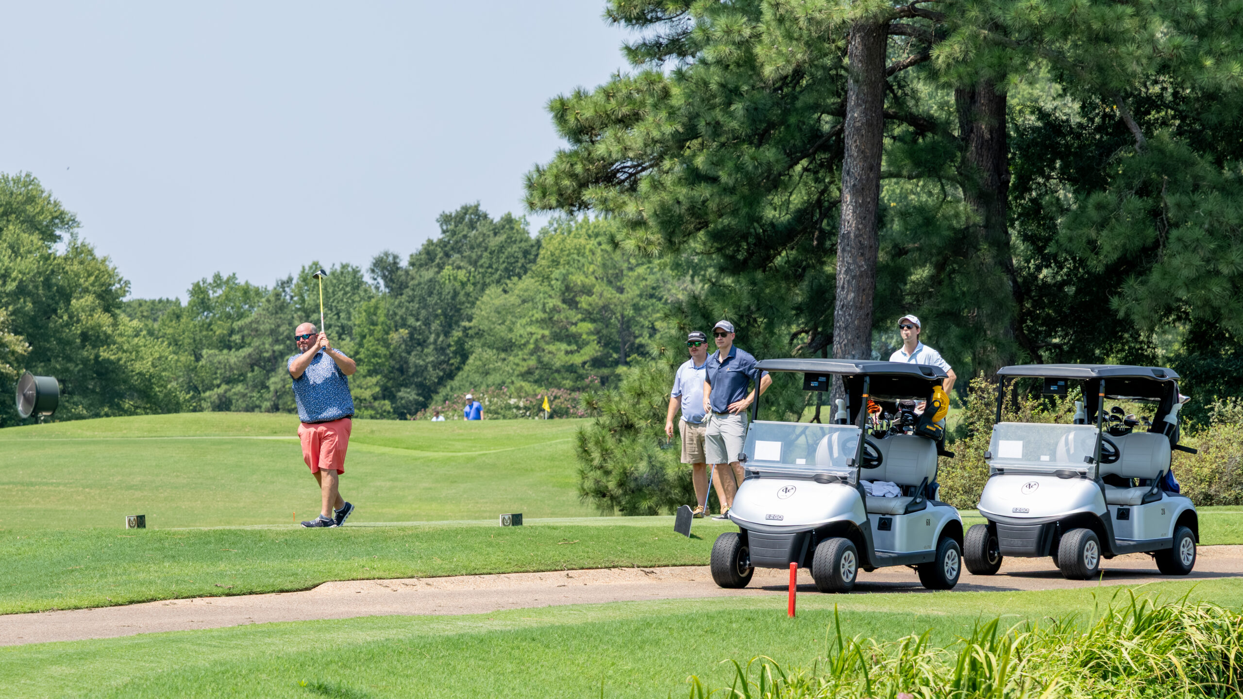 A golfer with an orange shirt and shorts stands on a tee box near two golf carts and others.