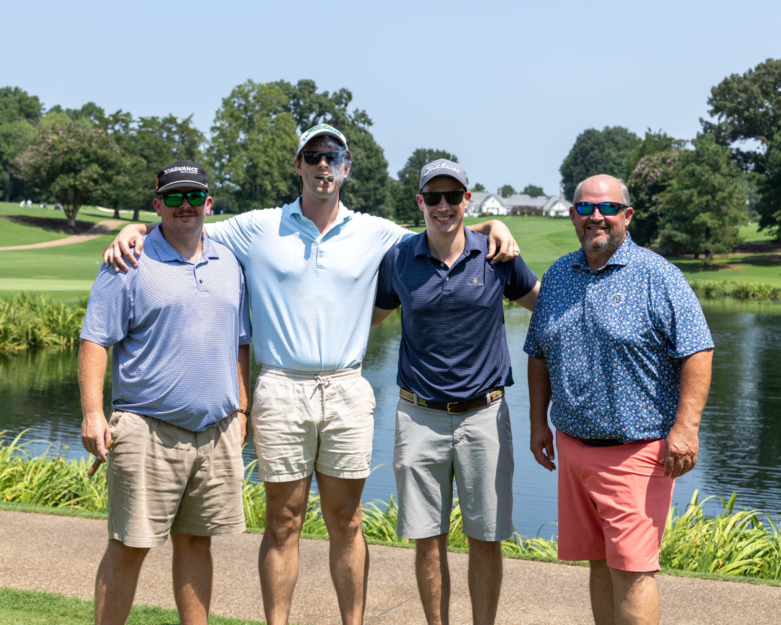 Four men in golf attire standing together on a path near a pond and golf course.