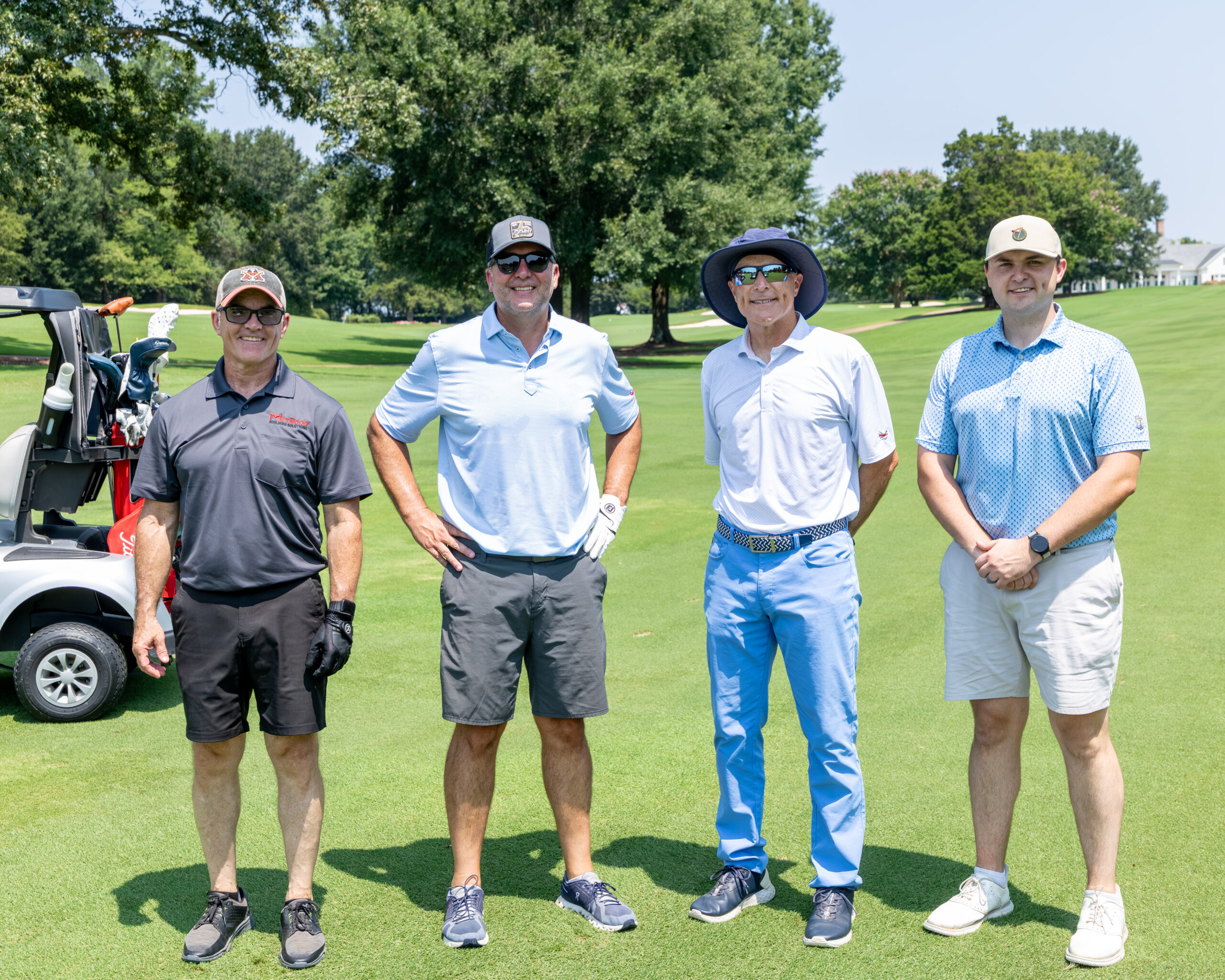 Four men standing together in front of a golf cart on a lush green grass field.