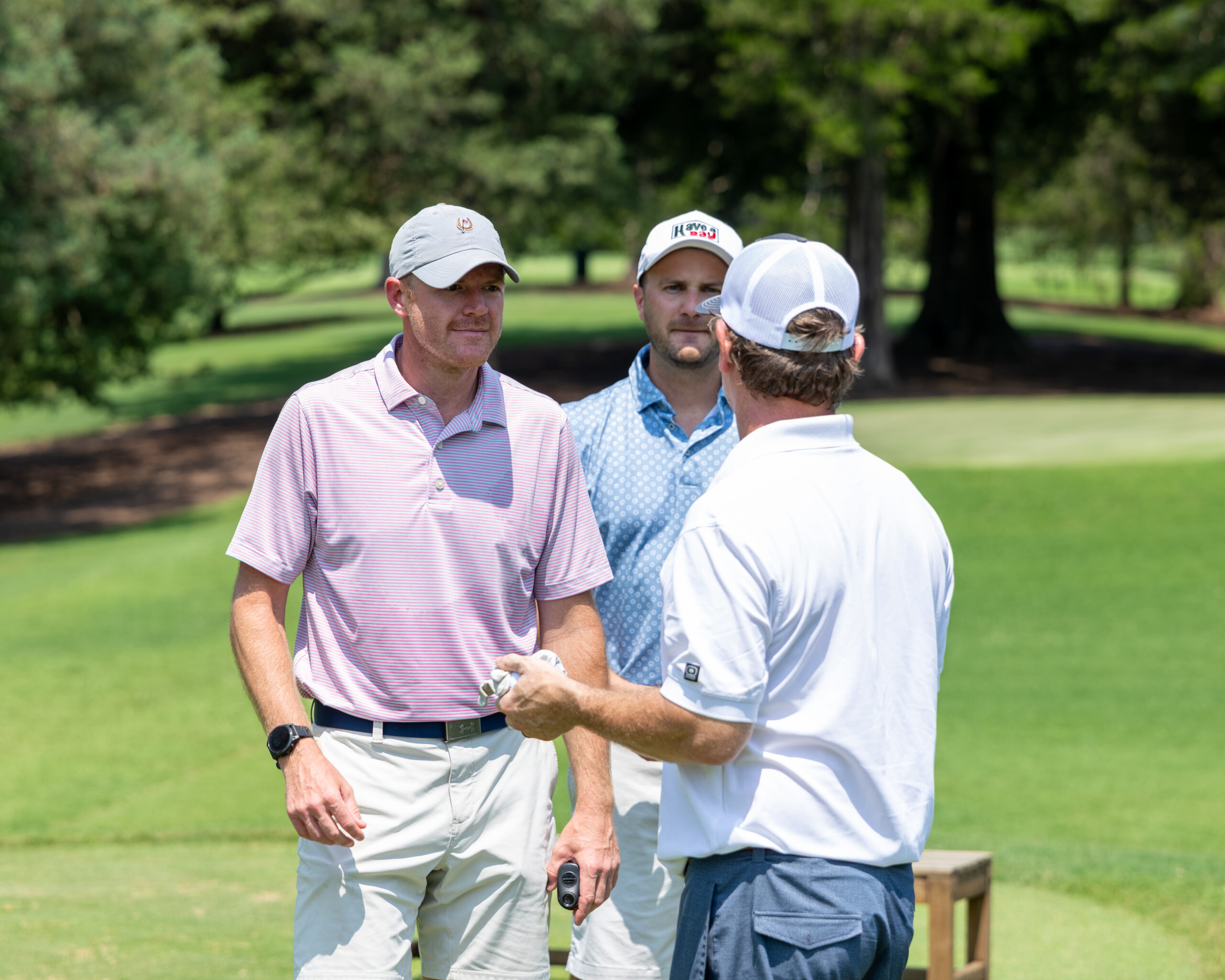 Three men in golf attire including hats and polos wear and interact on a green lawn during a tournament.