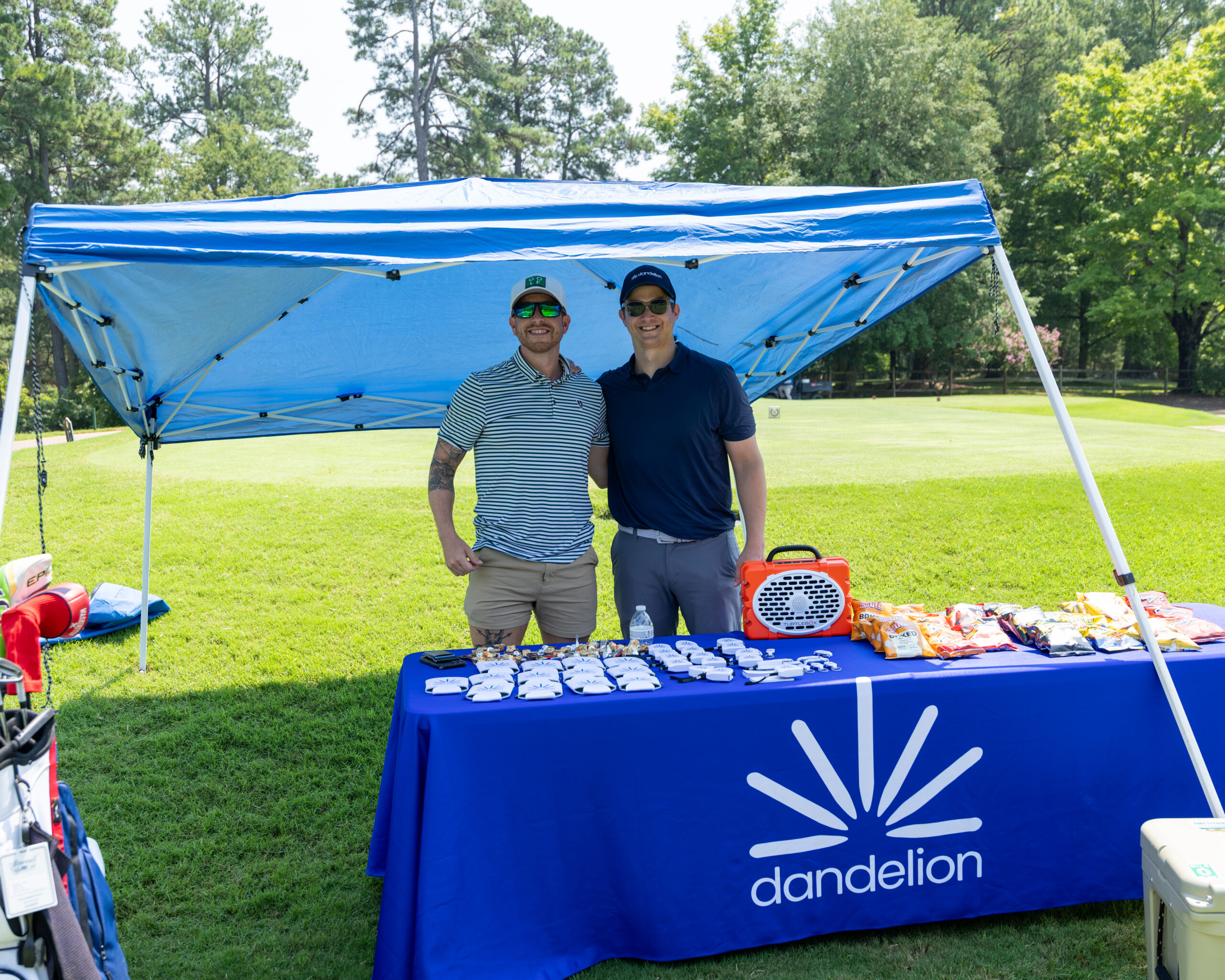 Two men standing behind a table with promotional items under a blue canopy tent.