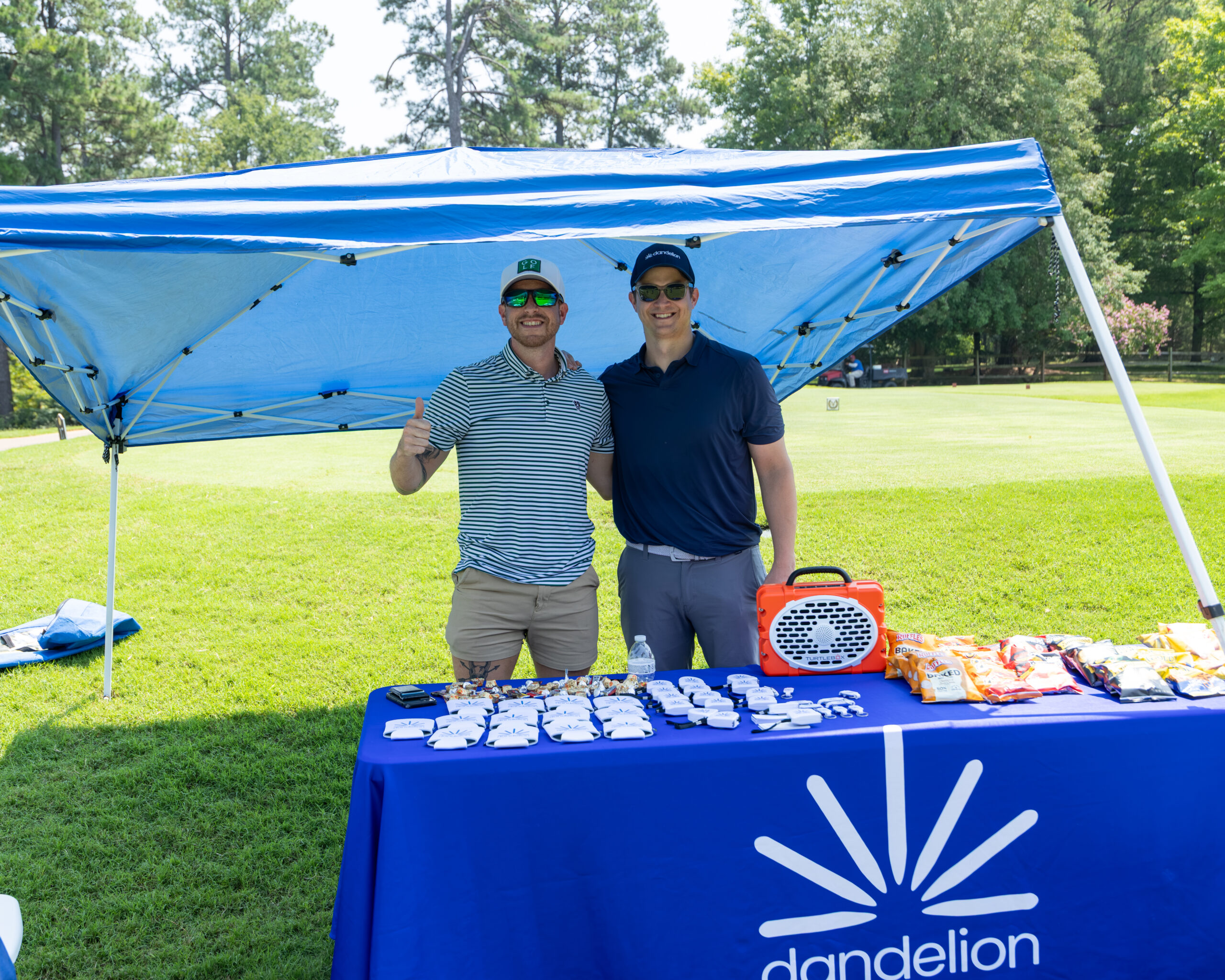 Two men standing behind a table with snacks and promotional items under a blue canopy at an outdoor golf course.