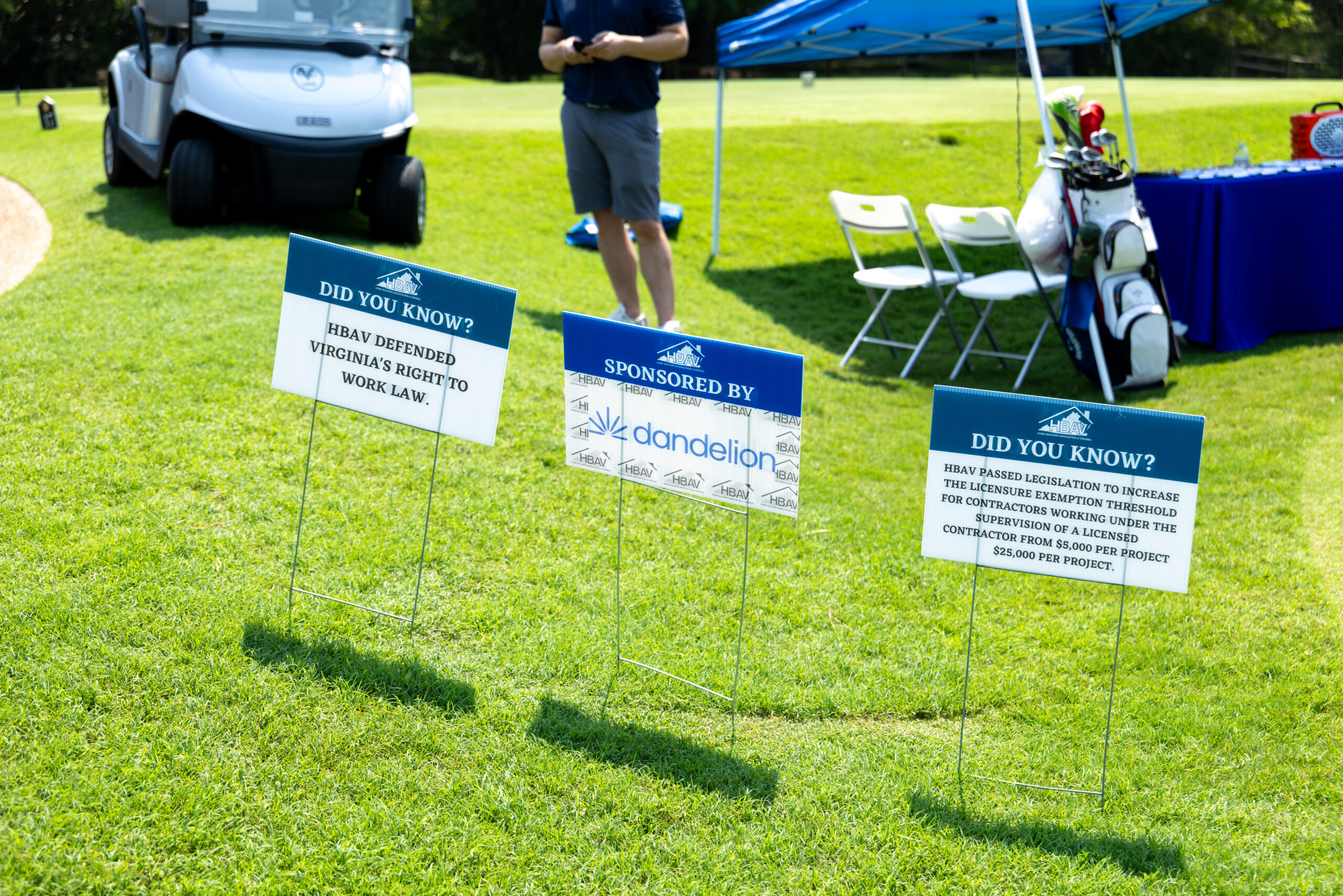 Two signs and a sponsor board are set up on a golf course with a golf cart and person in the background.