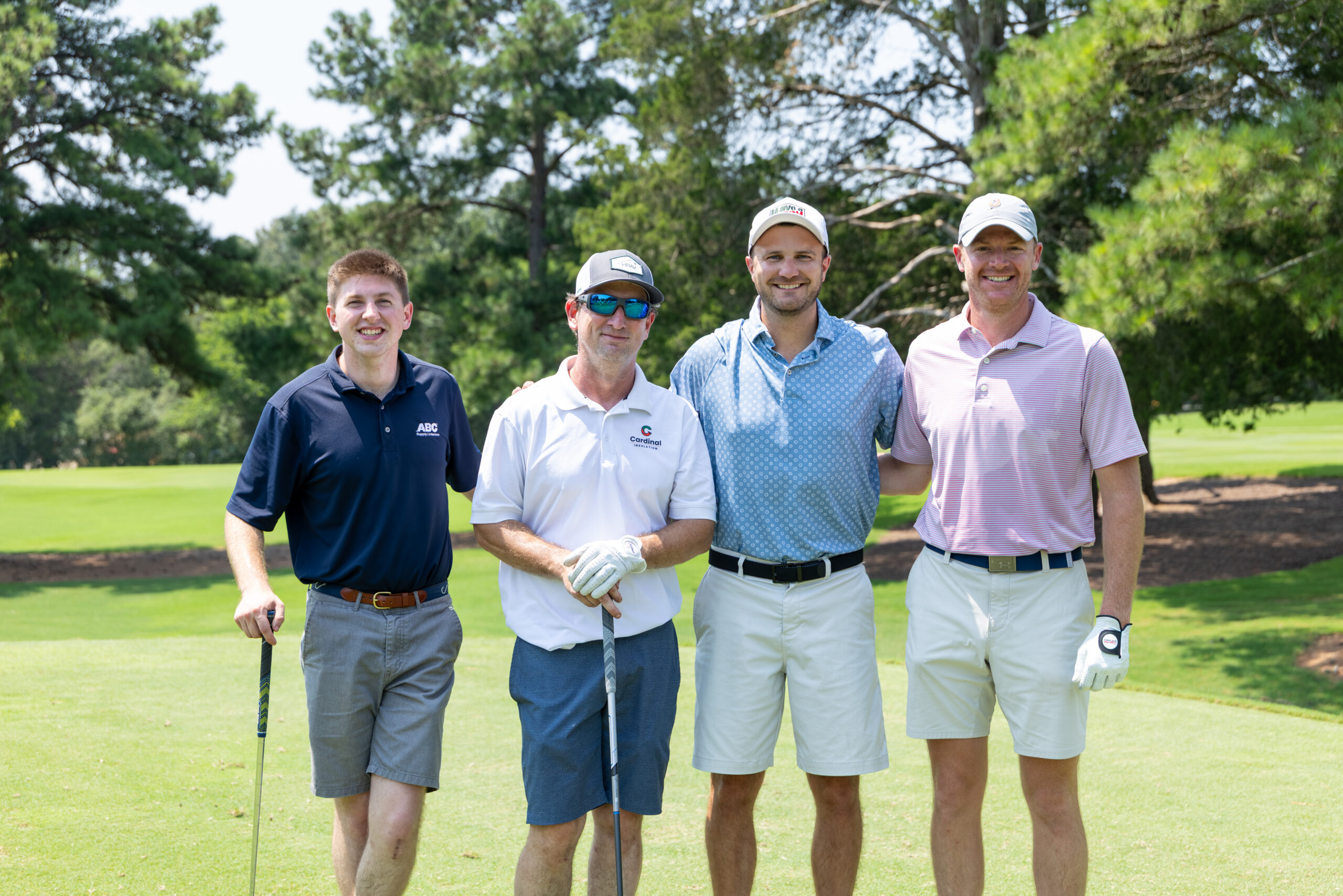 Four men standing and smiling for a photo on a green golf course with trees in the background.