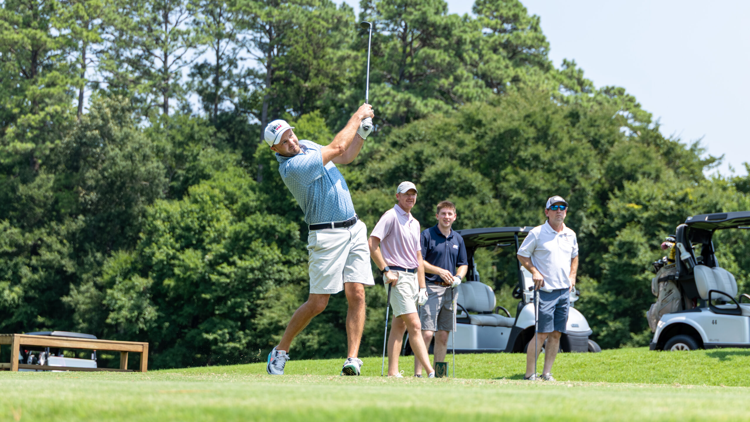 A golfer is taking a swing at a tee box on a course with three other men watching nearby.