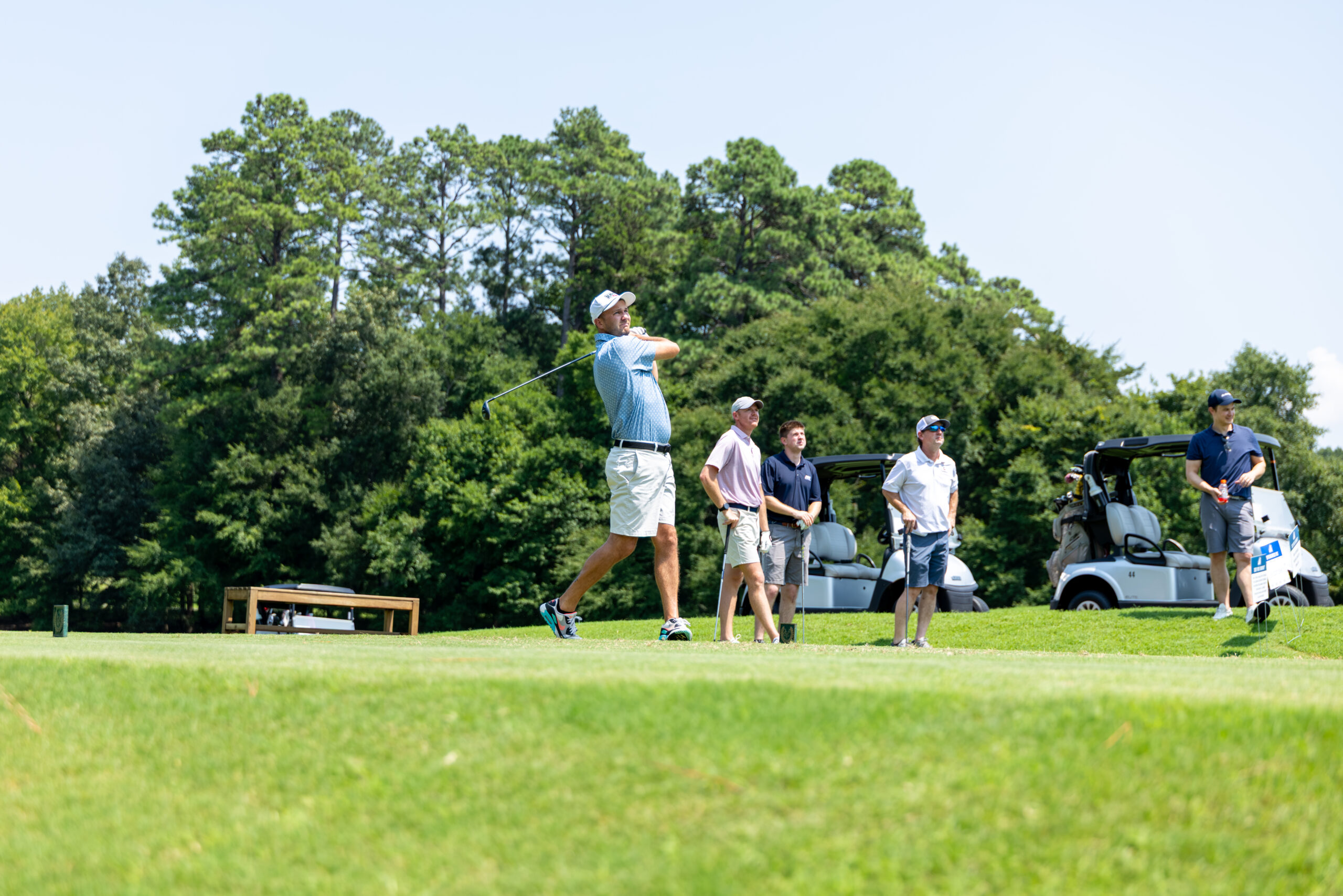 A golfer swings his club while others watch on the grass near golf carts.