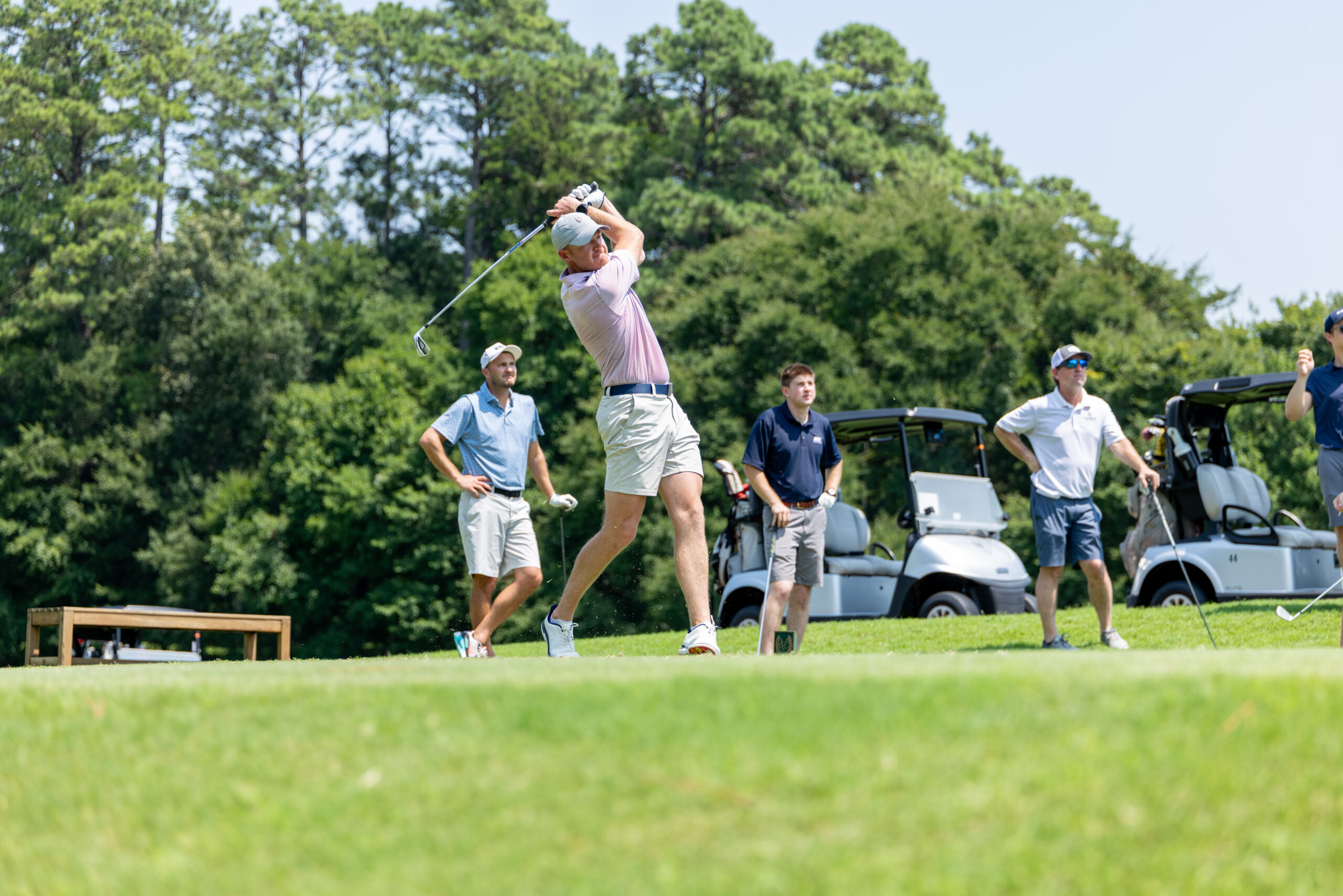 A golfer swings his club during a tournament on a golf course with others nearby.