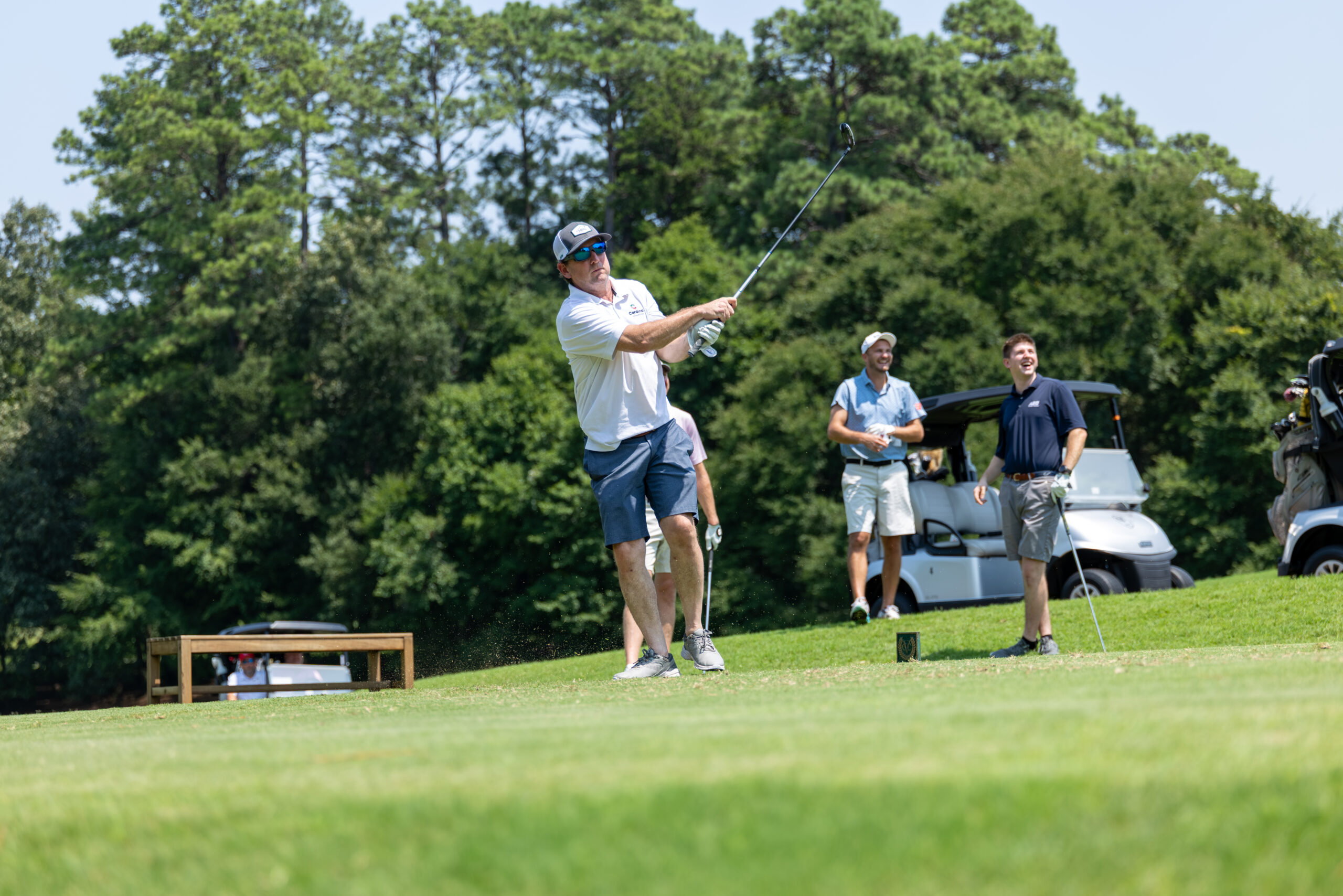 A man in white shirt and blue shorts kicks off his golf swing with others watching nearby on a grassy course.