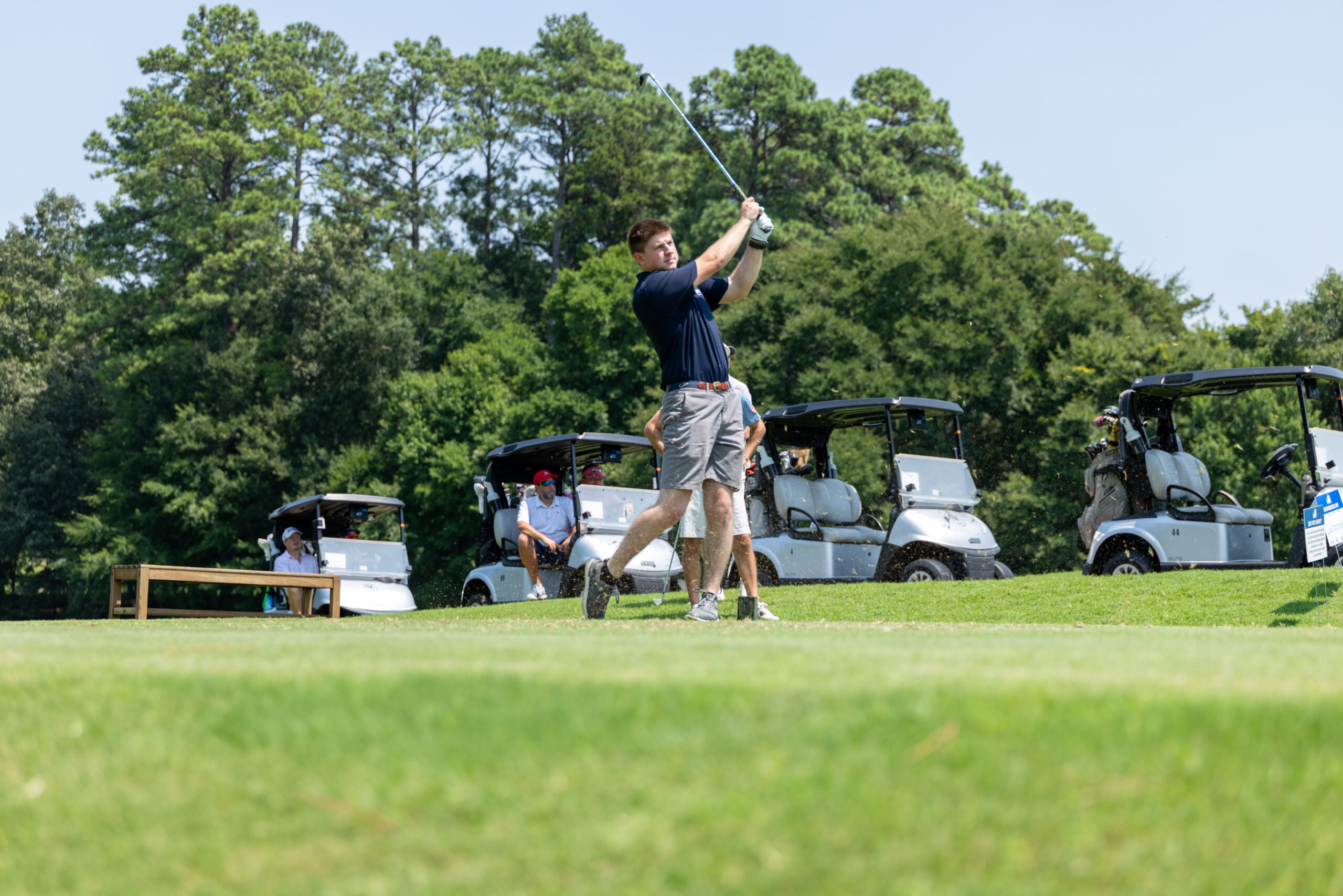 A man in a polo shirt and shorts golfs on a fairway with several carts and others watching.