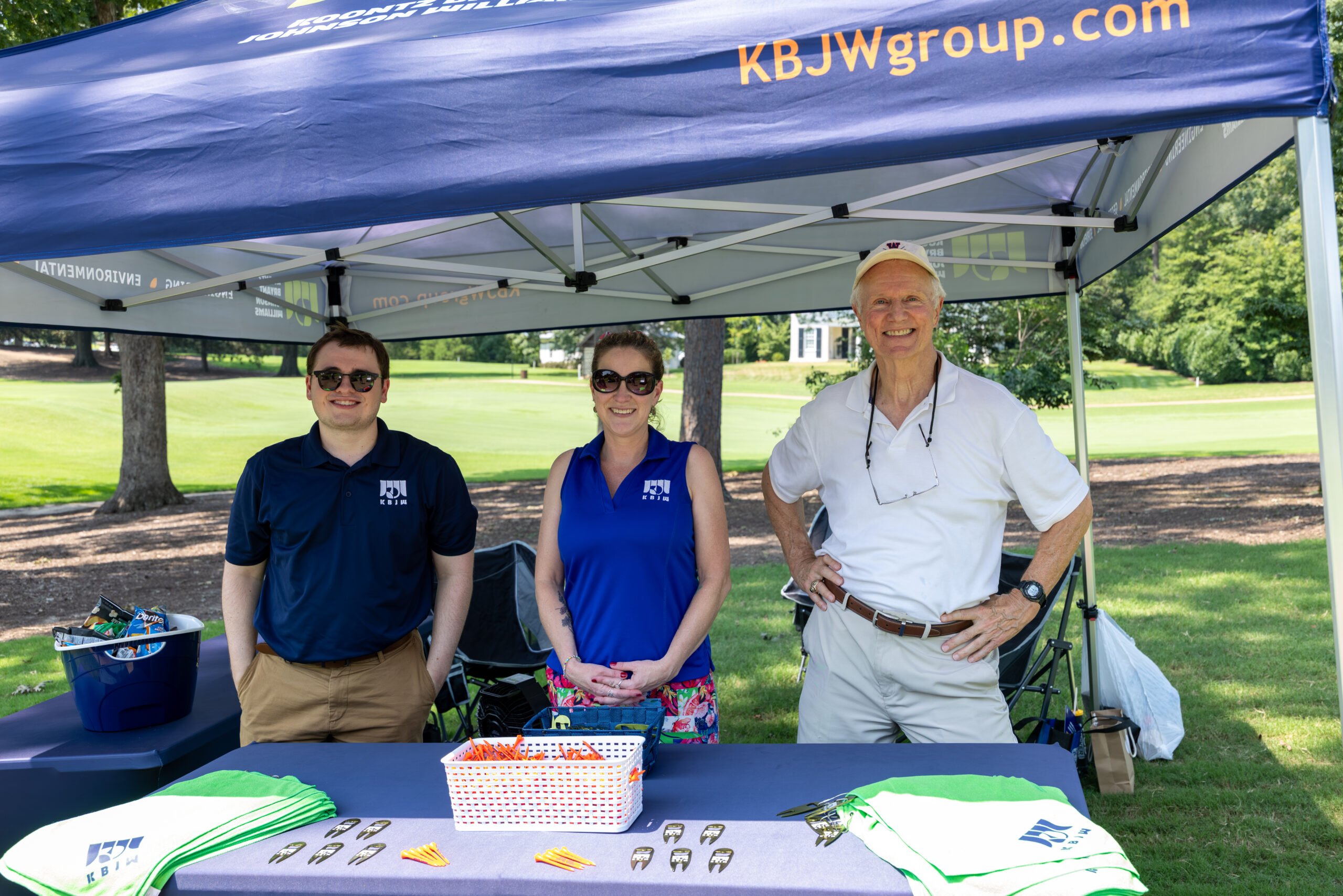 Three people standing under a blue canopy at a golf tournament booth.