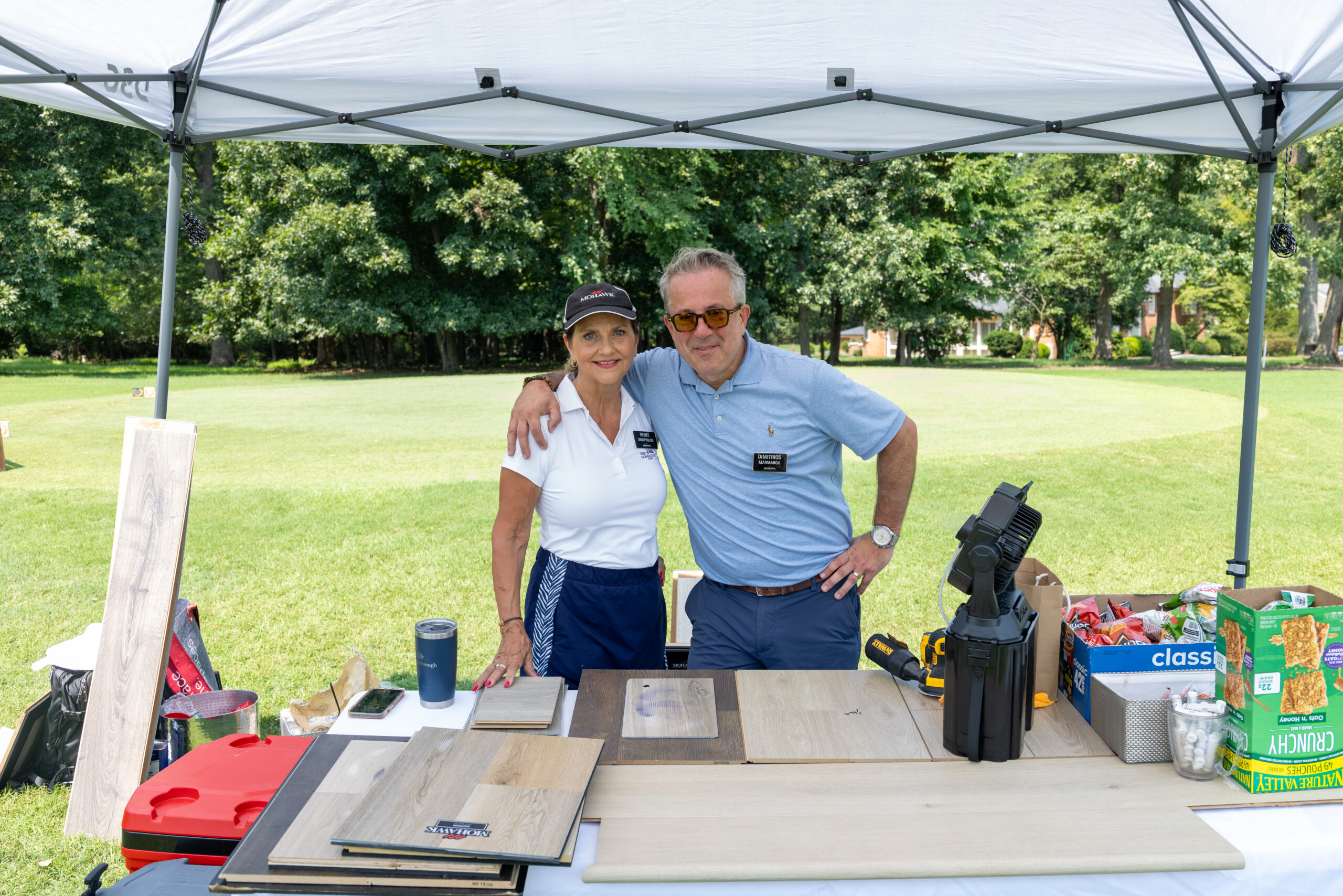 A man and woman stand behind a table filled with promotional items under a white tent at a golf course.