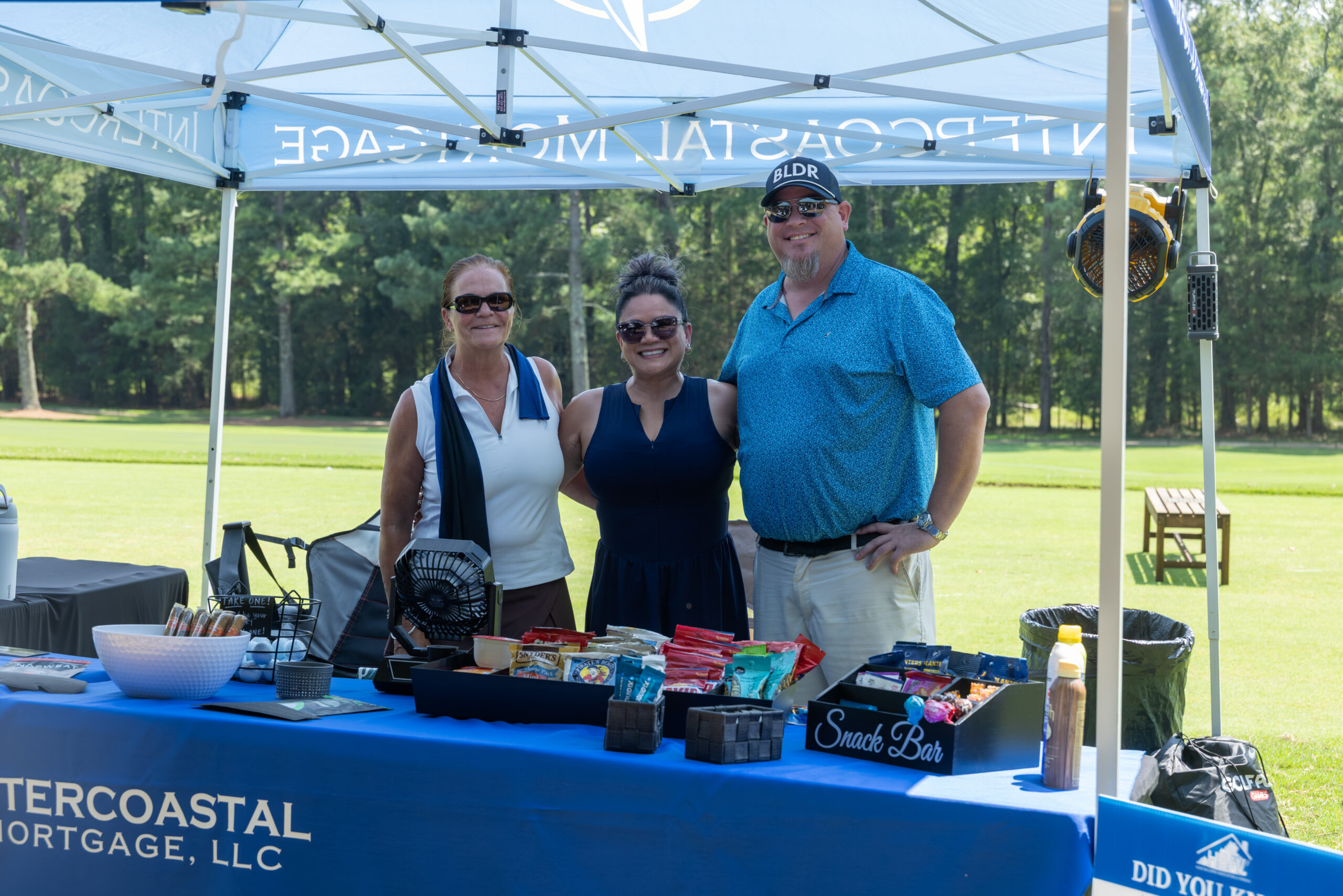Three people stand behind a table covered with snacks and promotional items under a branded canopy tent.