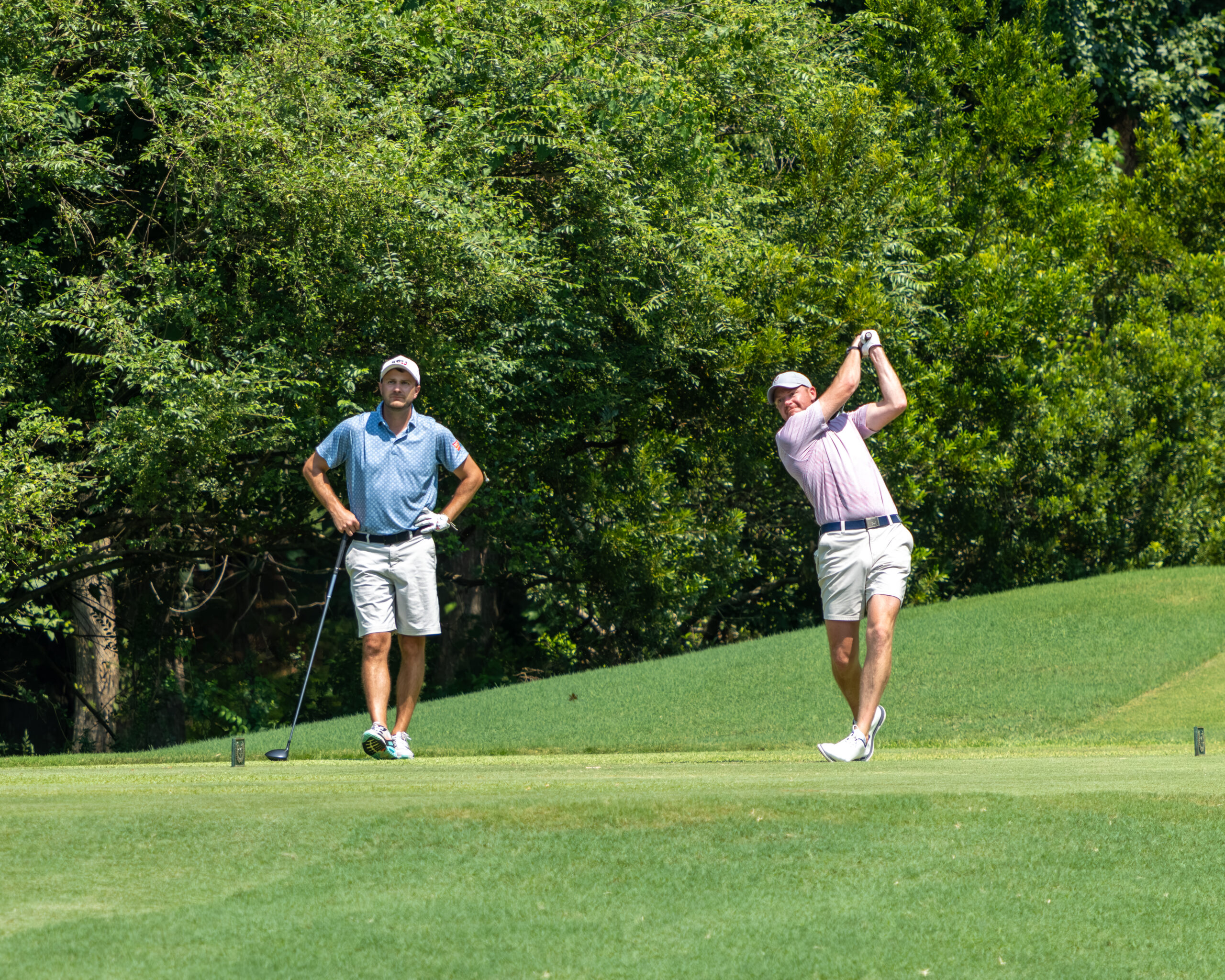 Two men on a golf course with one man and his golfing partner watch as another follows through after a swing.