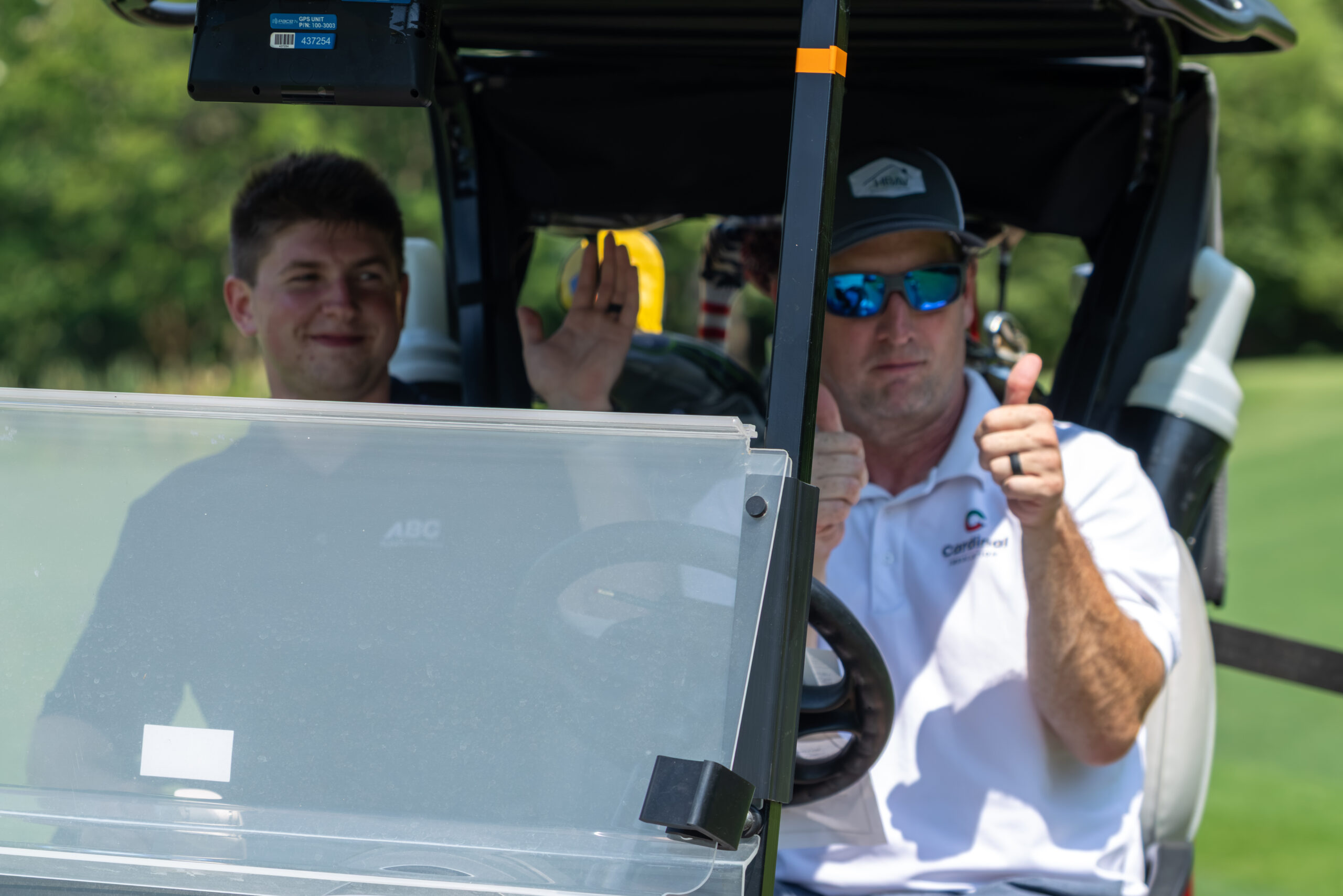 Two men on a golf cart carrying equipment, both giving thumbs up.