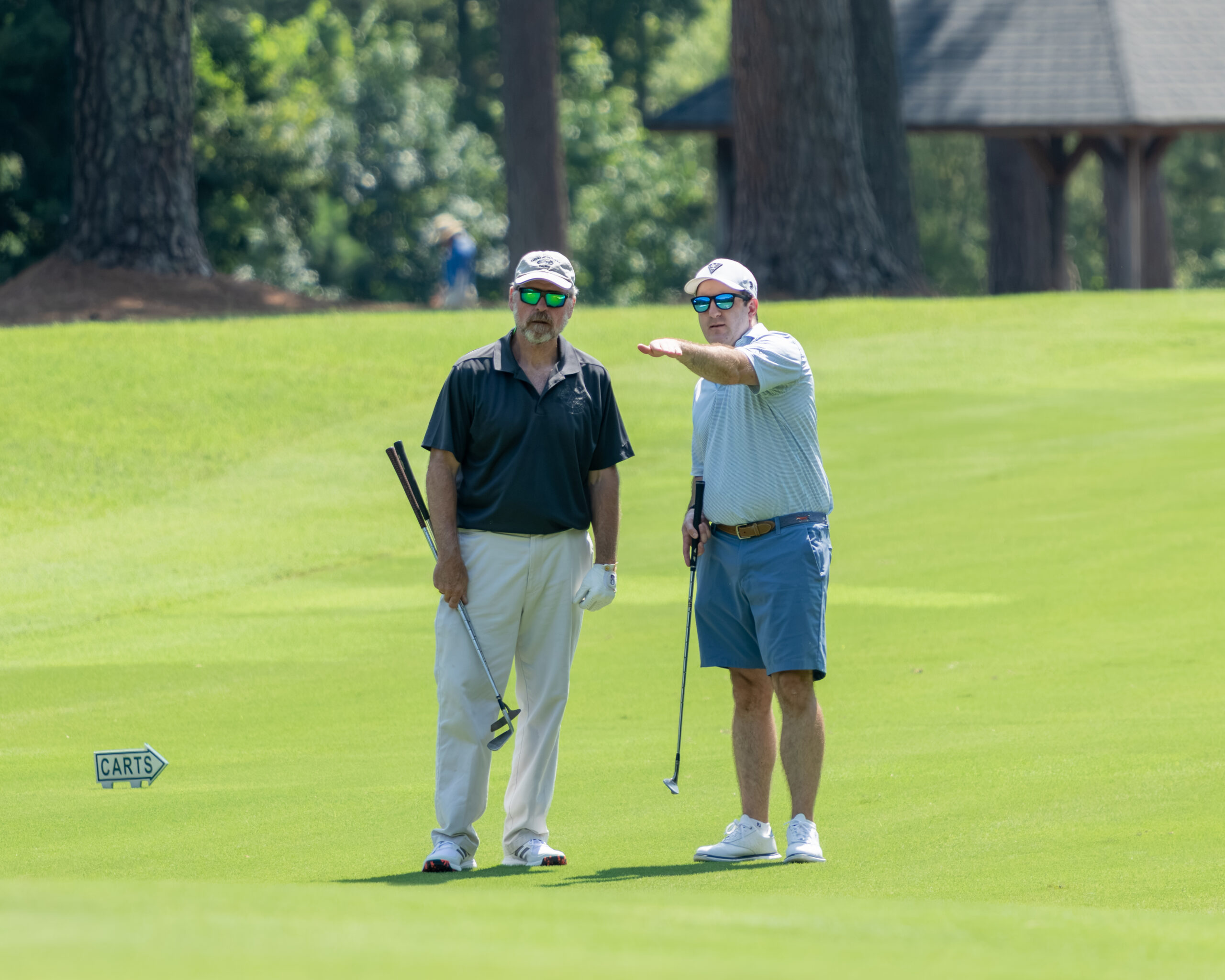 Two men on a golf course green near tall trees and a building.