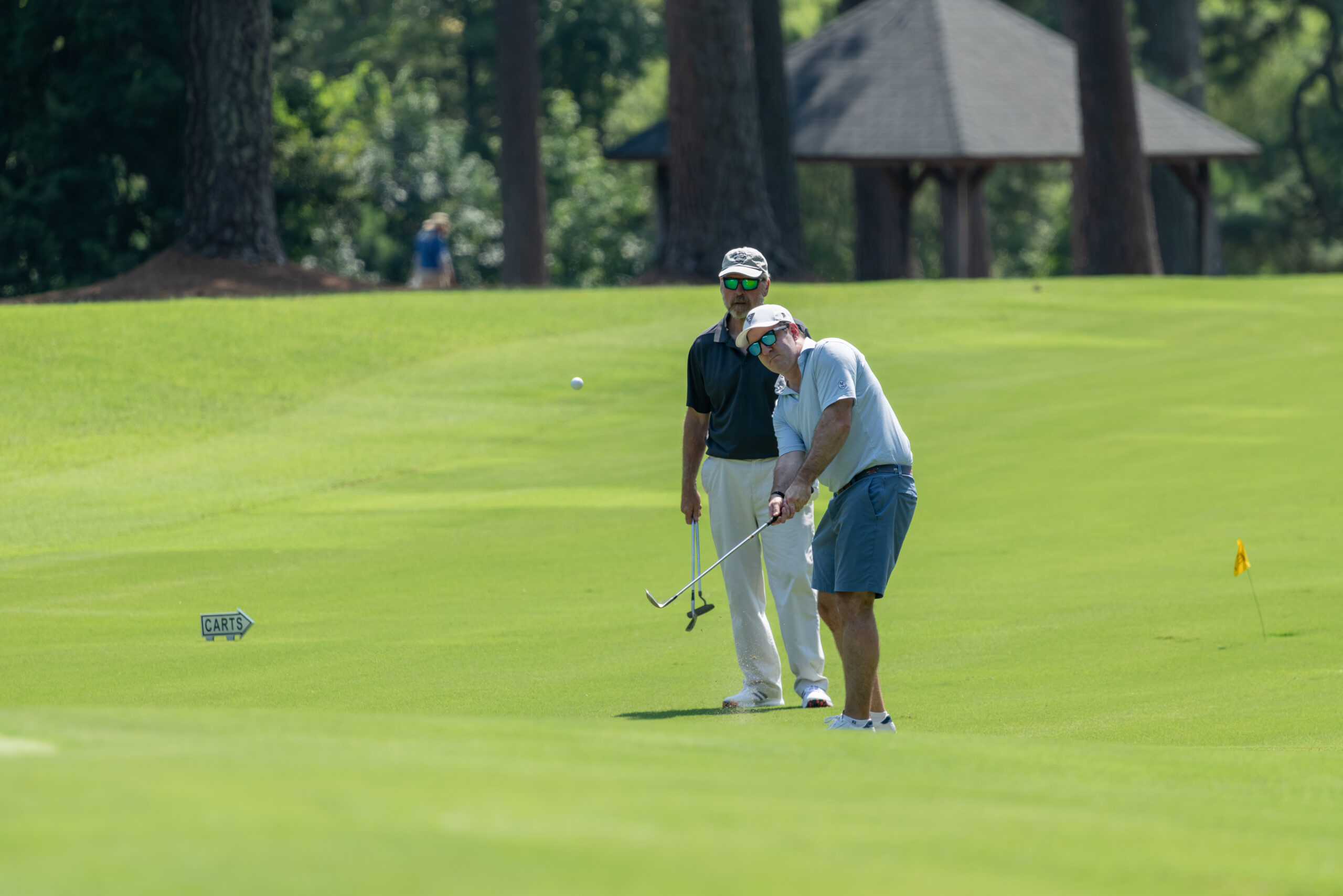 Two men on a golf green, one putting with a club and another watching from behind.