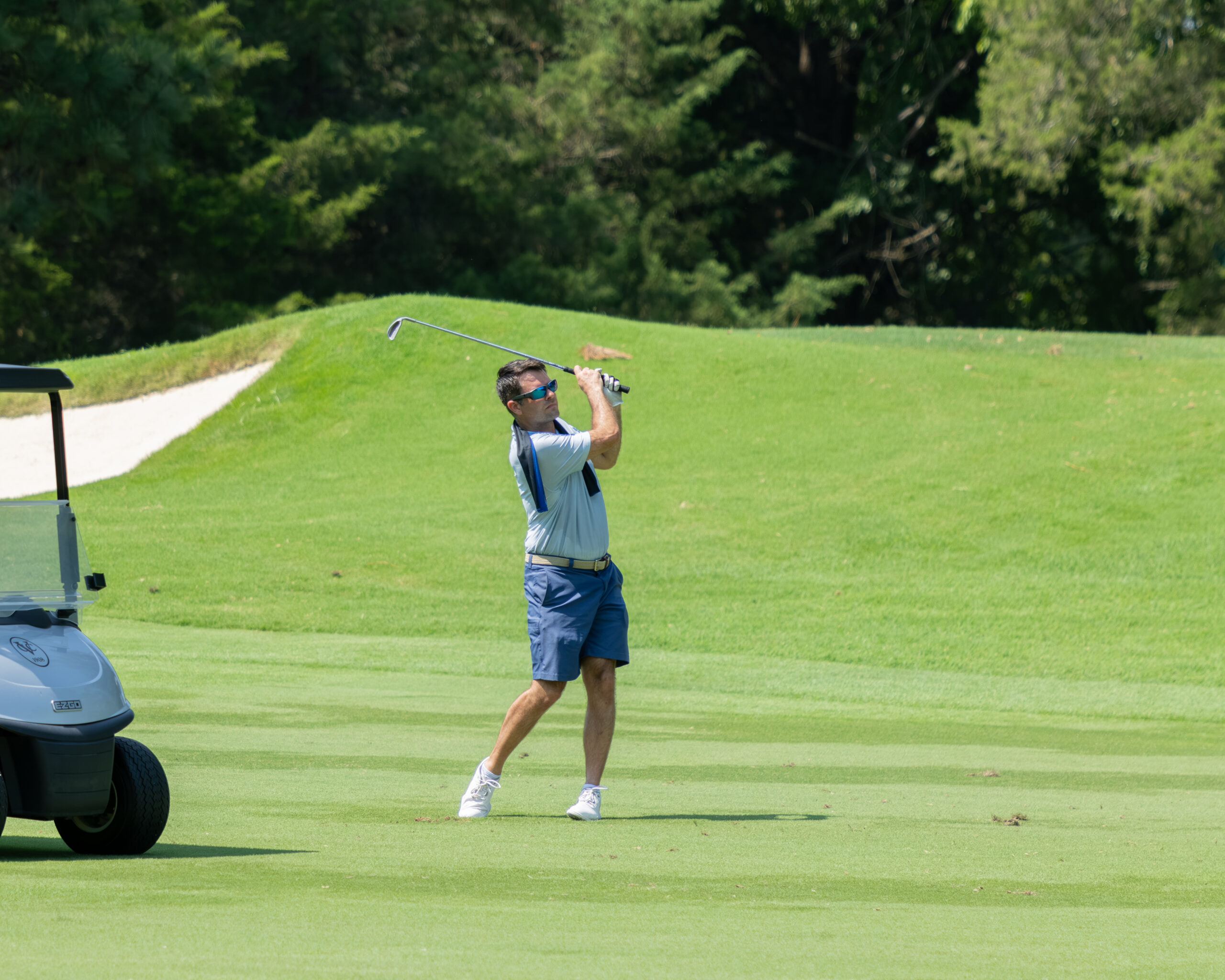 A man golfer in a blue shirt and shorts is taking a swing with a club on a green grass field.