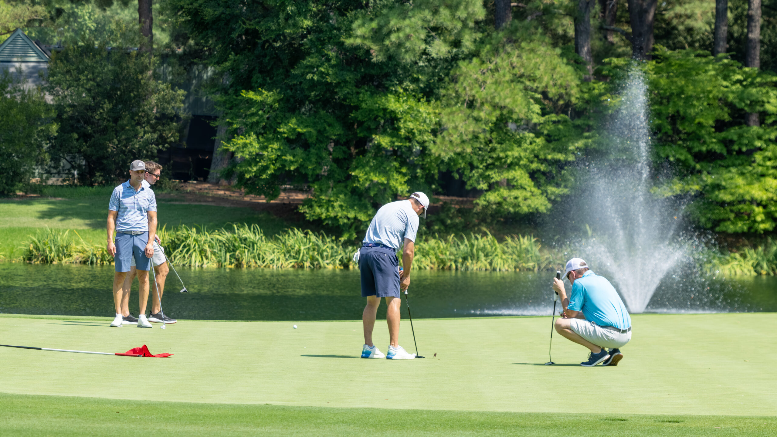 Three men on a green while playing golf near a fountain and water hazard.