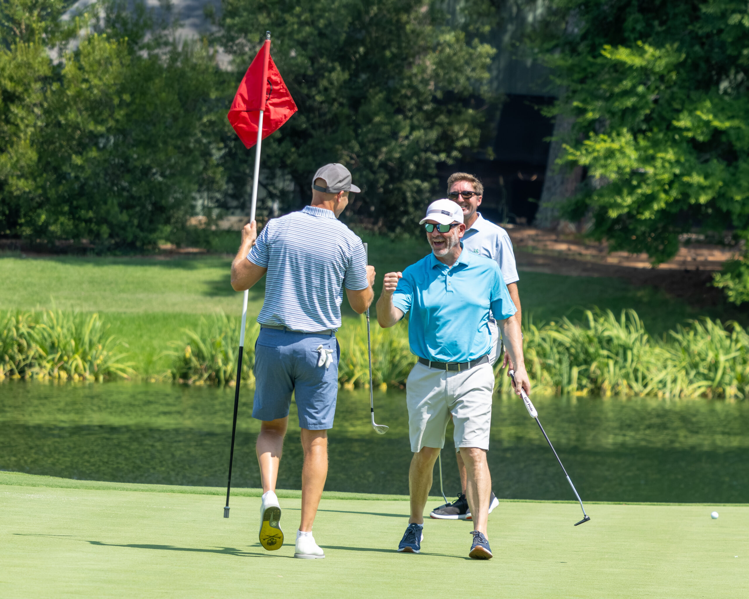 Three golfers on a green with a red flag, one man cheering and another holding a club.
