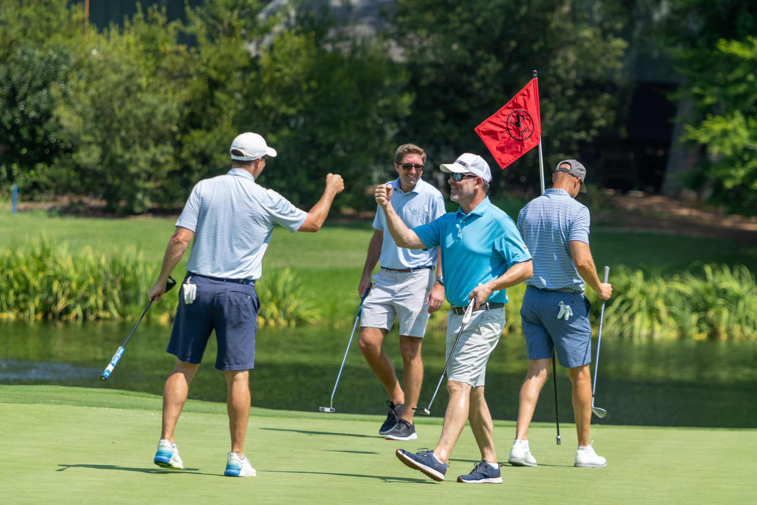Four men on a golf green near a red flag with their golf clubs and putters.