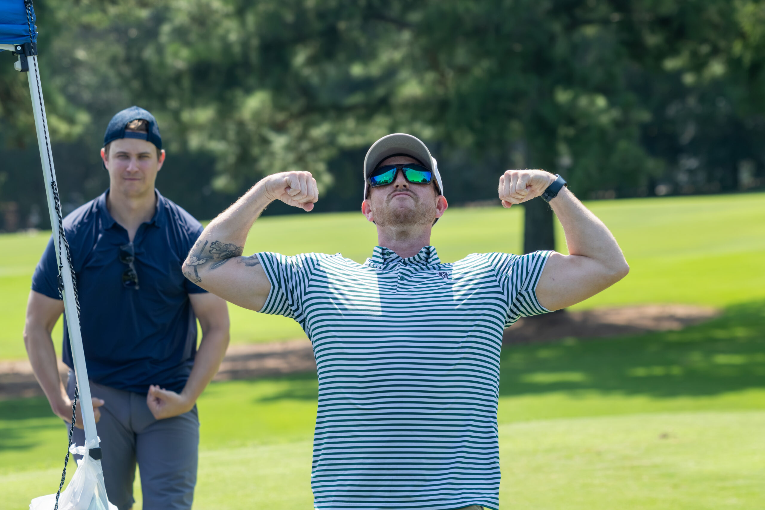 A man in a striped shirt and sunglasses flexes his muscles while another man watches in the background.