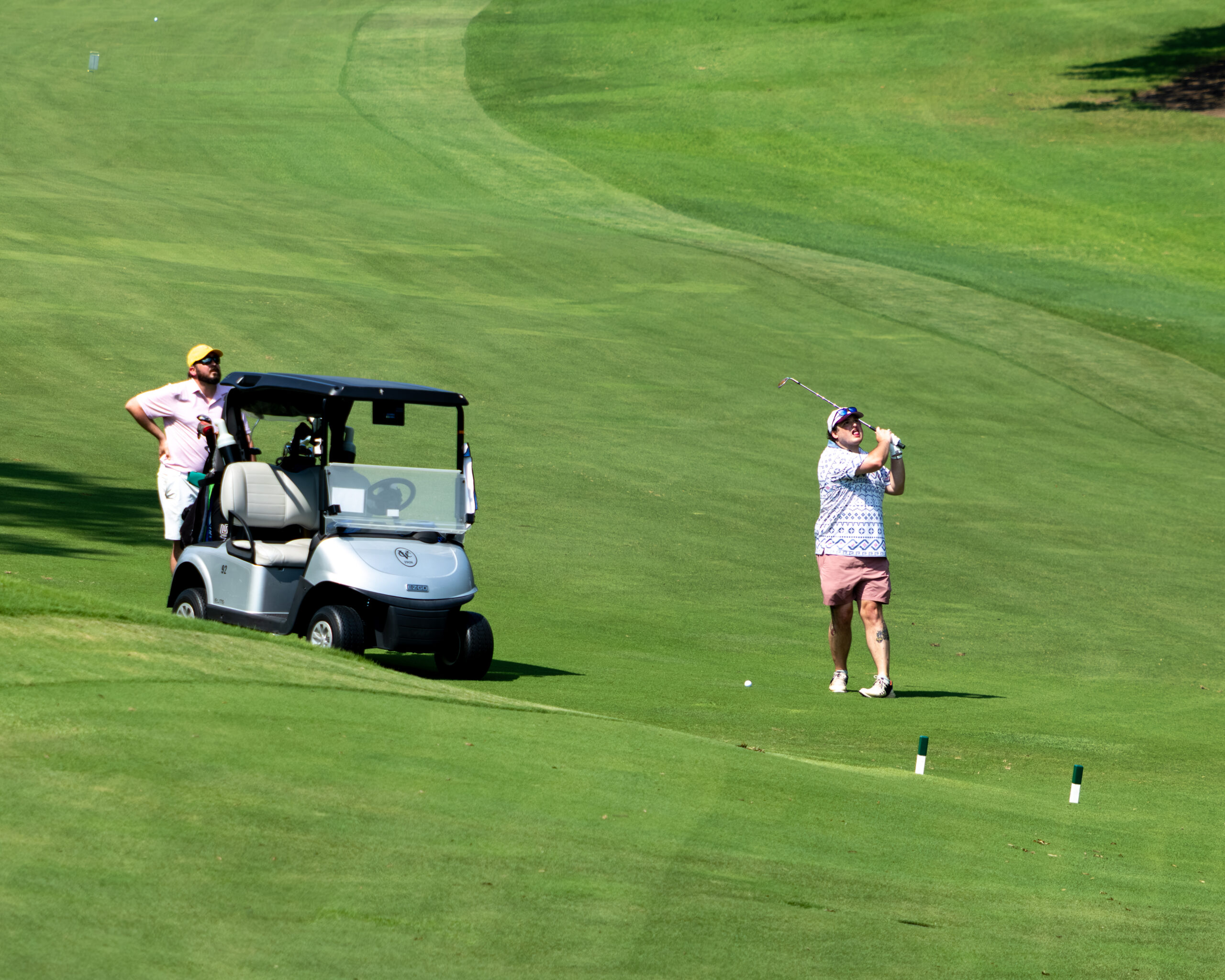 A golfer wearing sunglasses and a patterned shirt stands on a fairway with a golf club over his head.
