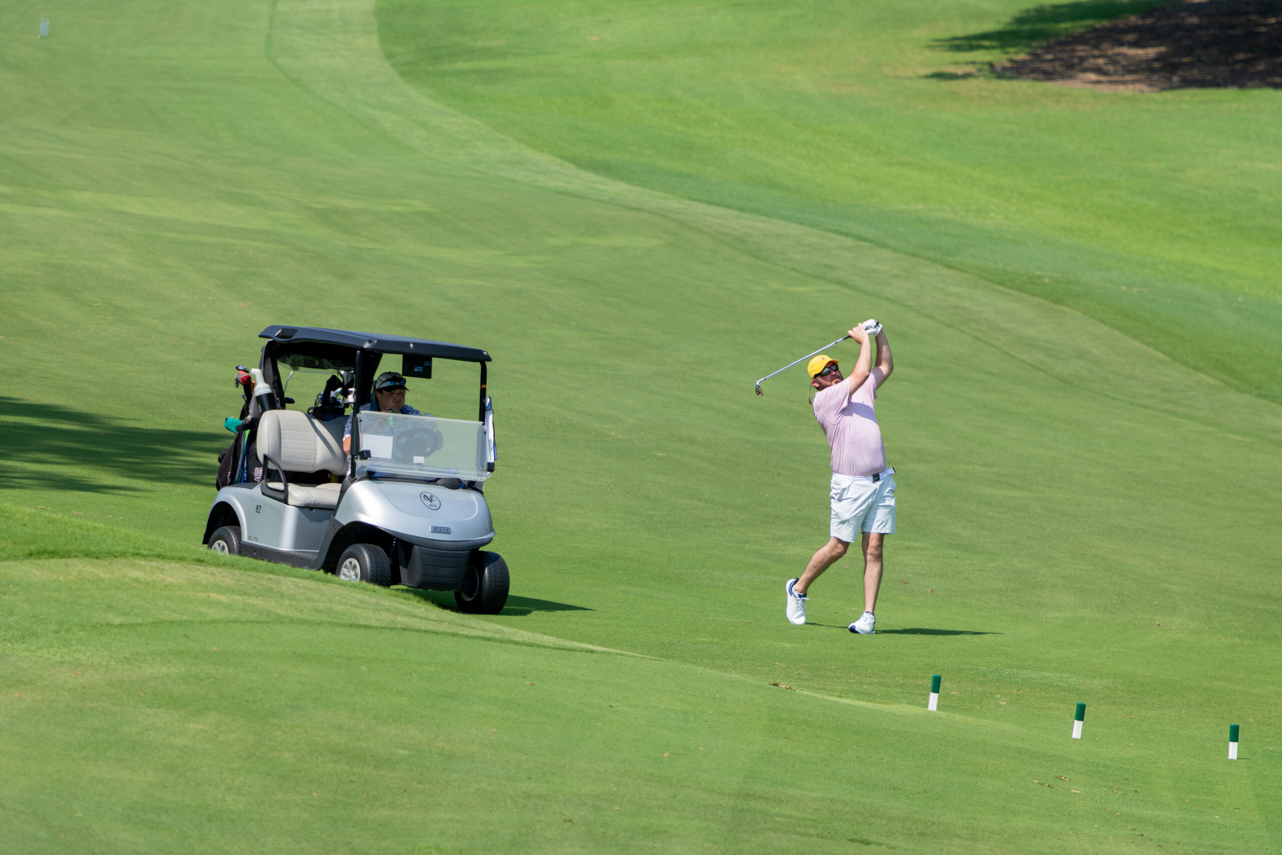 A golfer in a pink shirt and white shorts swings at a ball on a green grass course with a golf cart nearby.
