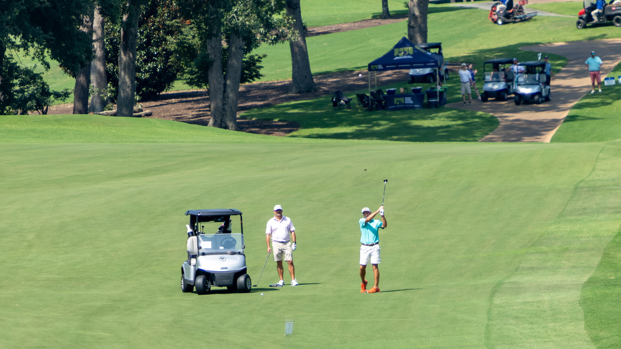 Two men on a golf course green, one putting and another standing nearby with a golf cart.