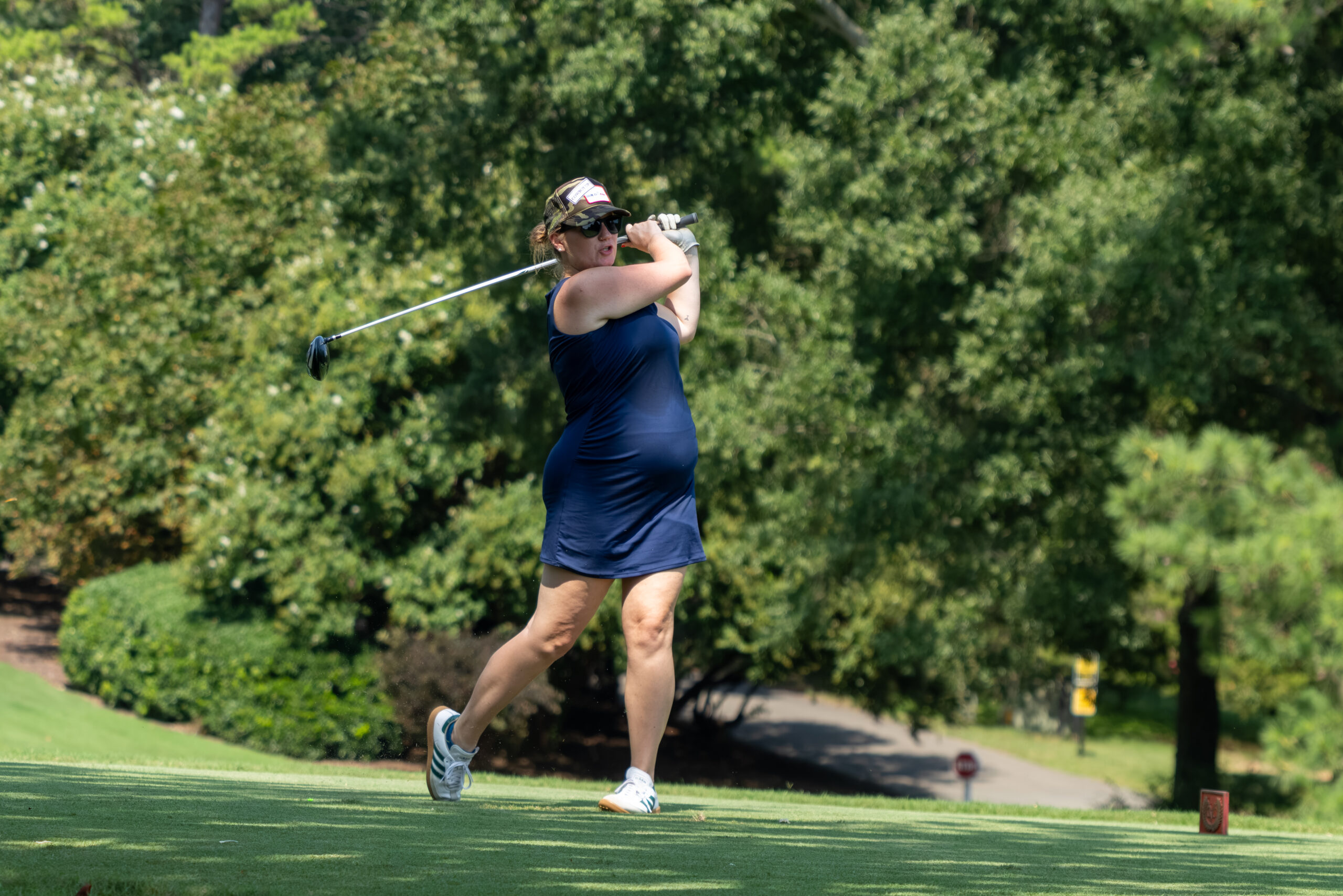A woman golfer in a blue dress and camo hat leans back as she completes her swing.