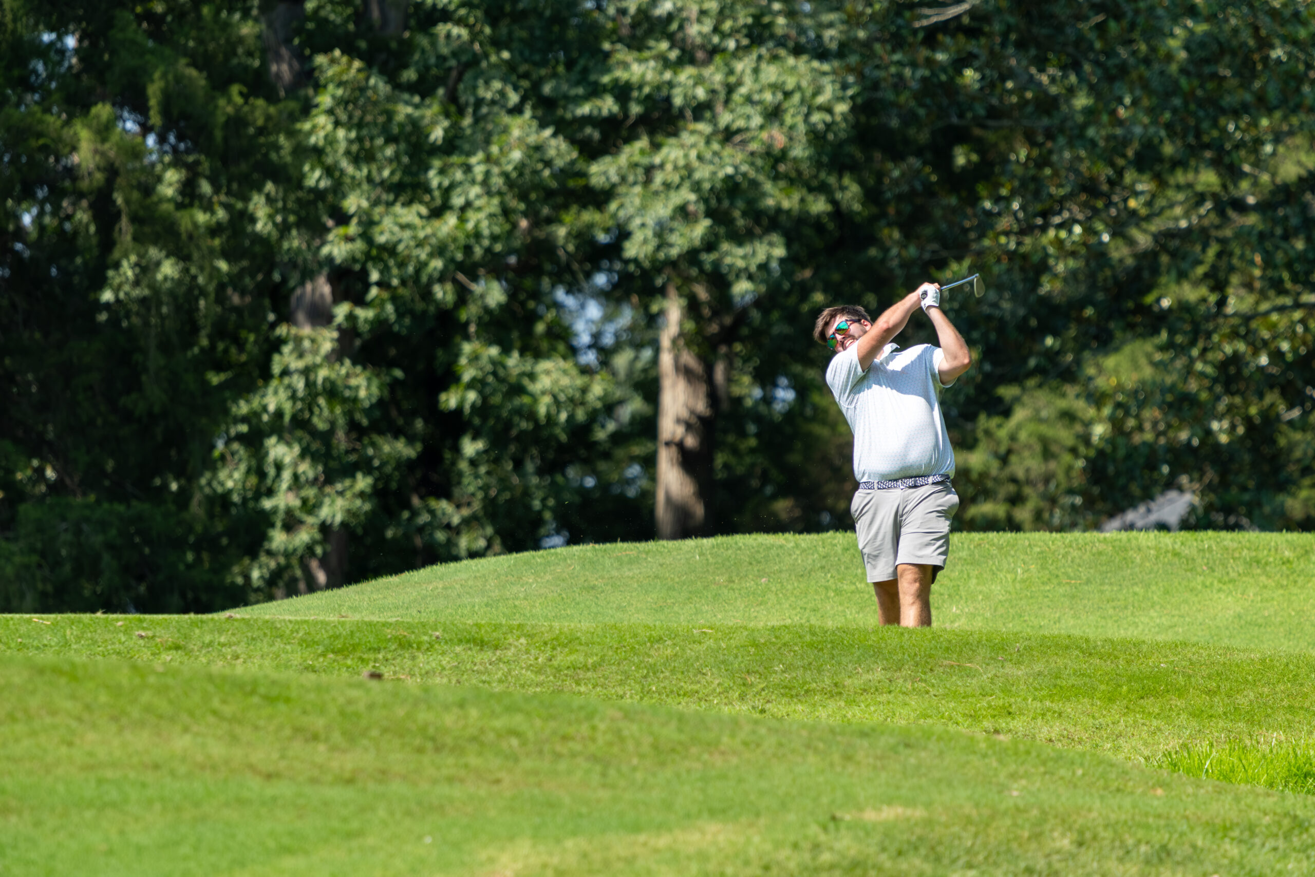 A golfer in a blue shirt and khaki shorts is swinging a golf club on a green field near trees.