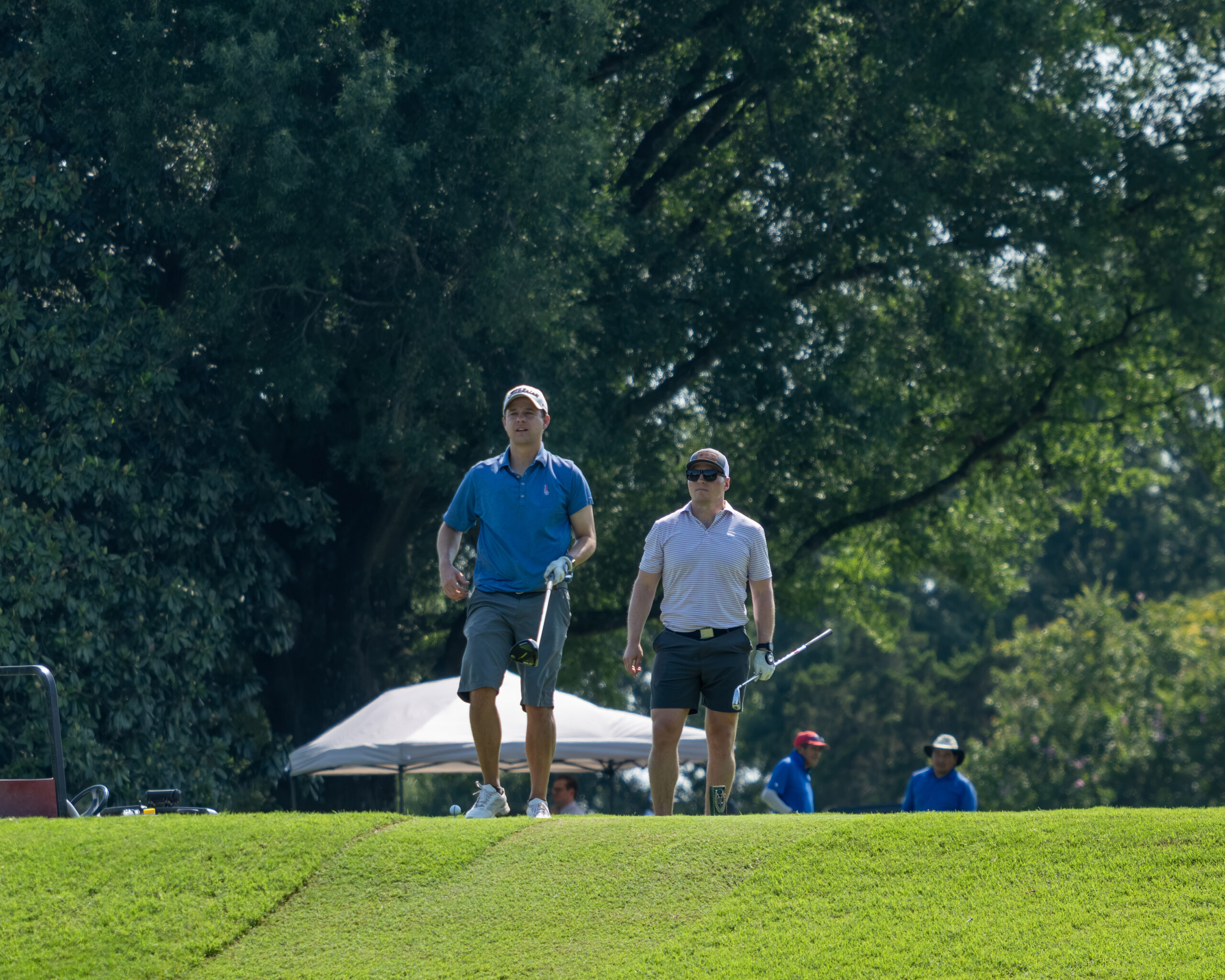 Two men golfing on a green near trees and a white tent under bright sunlight.