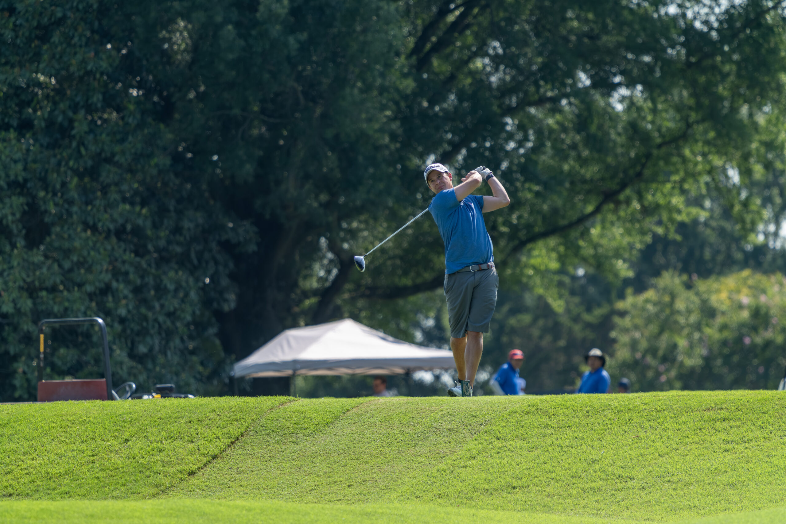 A male golfer in a blue shirt and grey shorts is following through after a golf swing on a grassy mound.