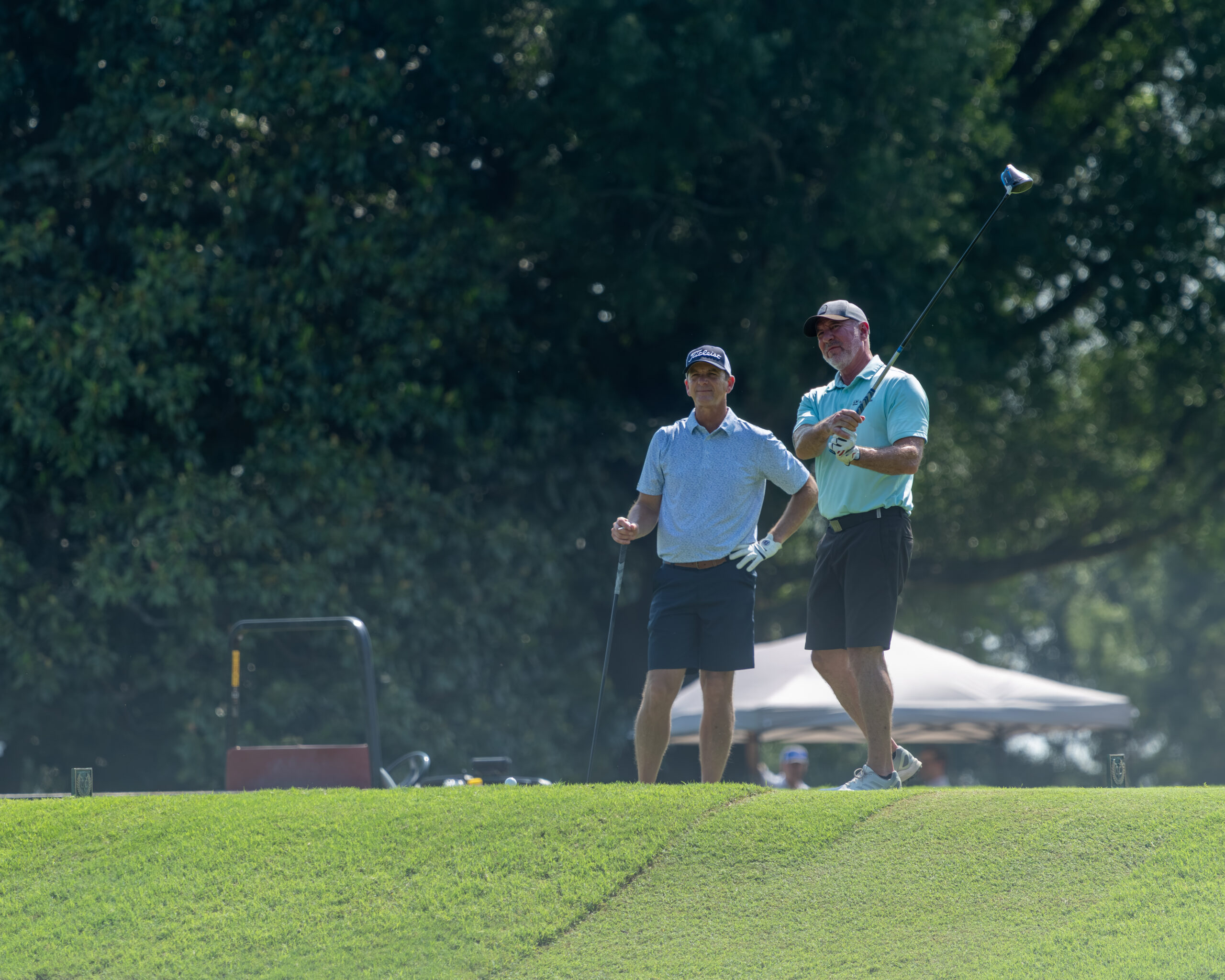Two men in golf attire stand on a green and hold clubs, looking towards each other.
