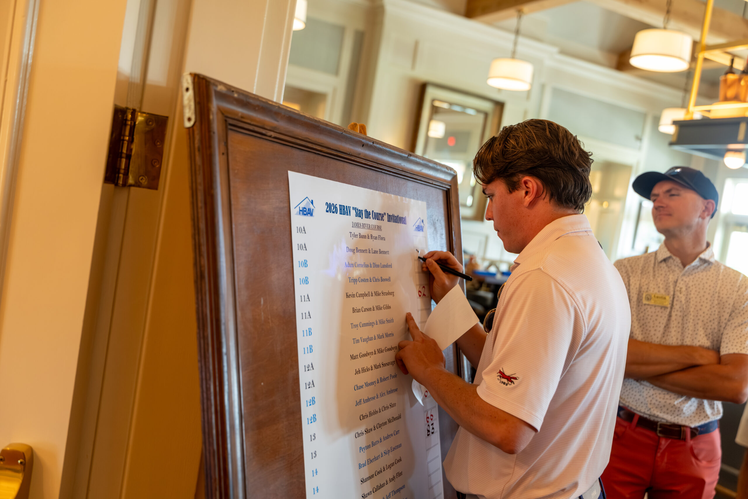 A man in a white polo shirt at the leaderboard on a at a golf tournament.