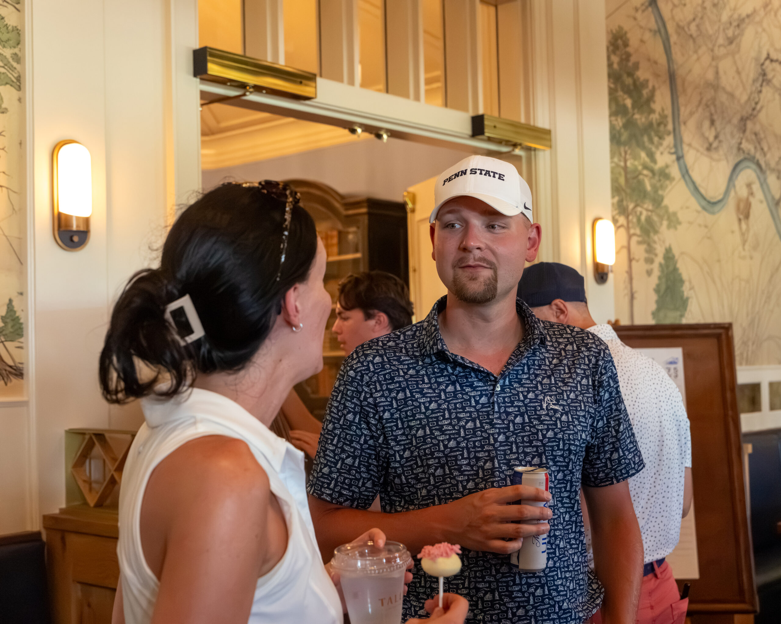 A woman and a man wearing a baseball cap hold drinks while talking in an indoor venue.