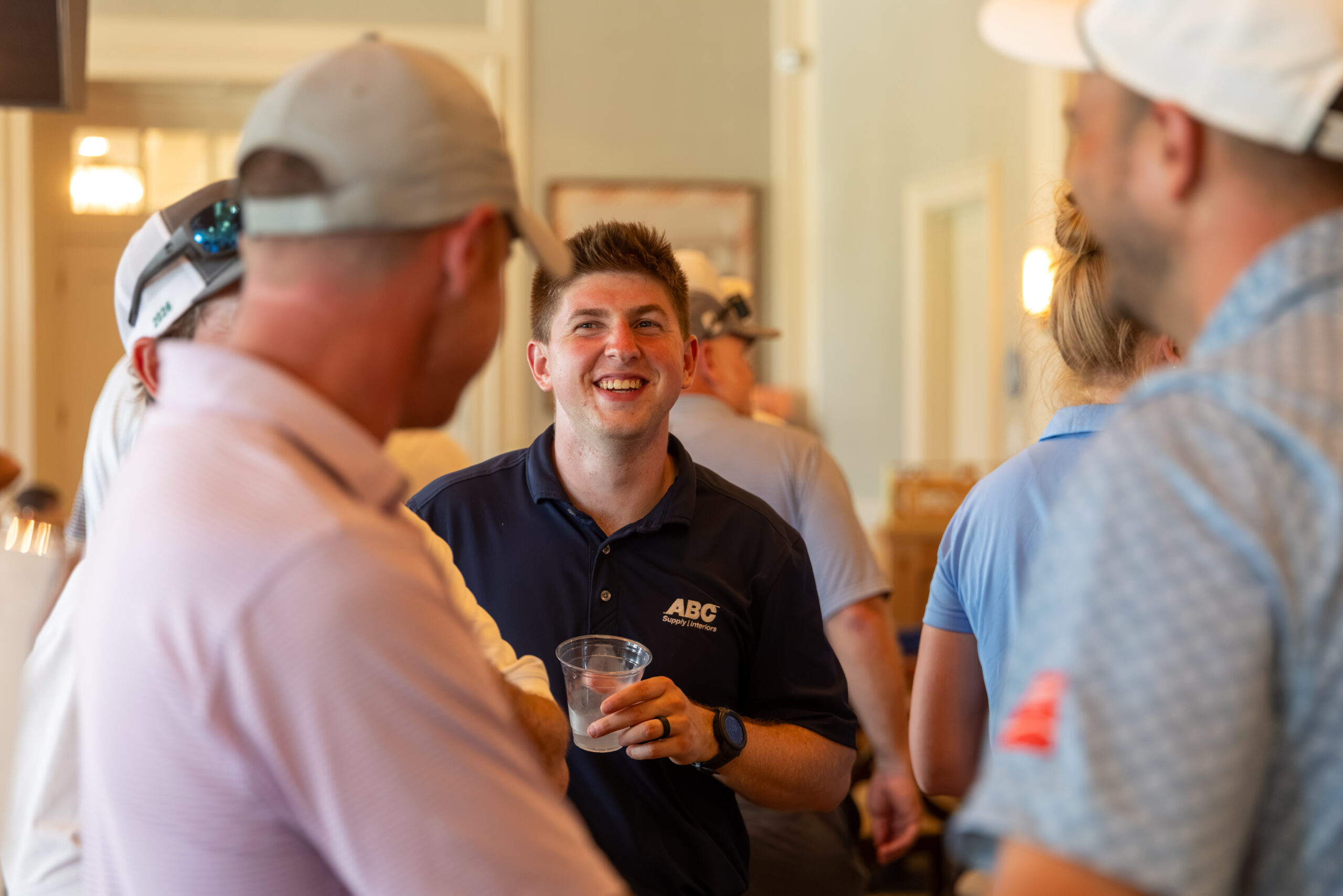 A man in a blue polo shirt smiles while holding a drink and talking to others in a crowd.