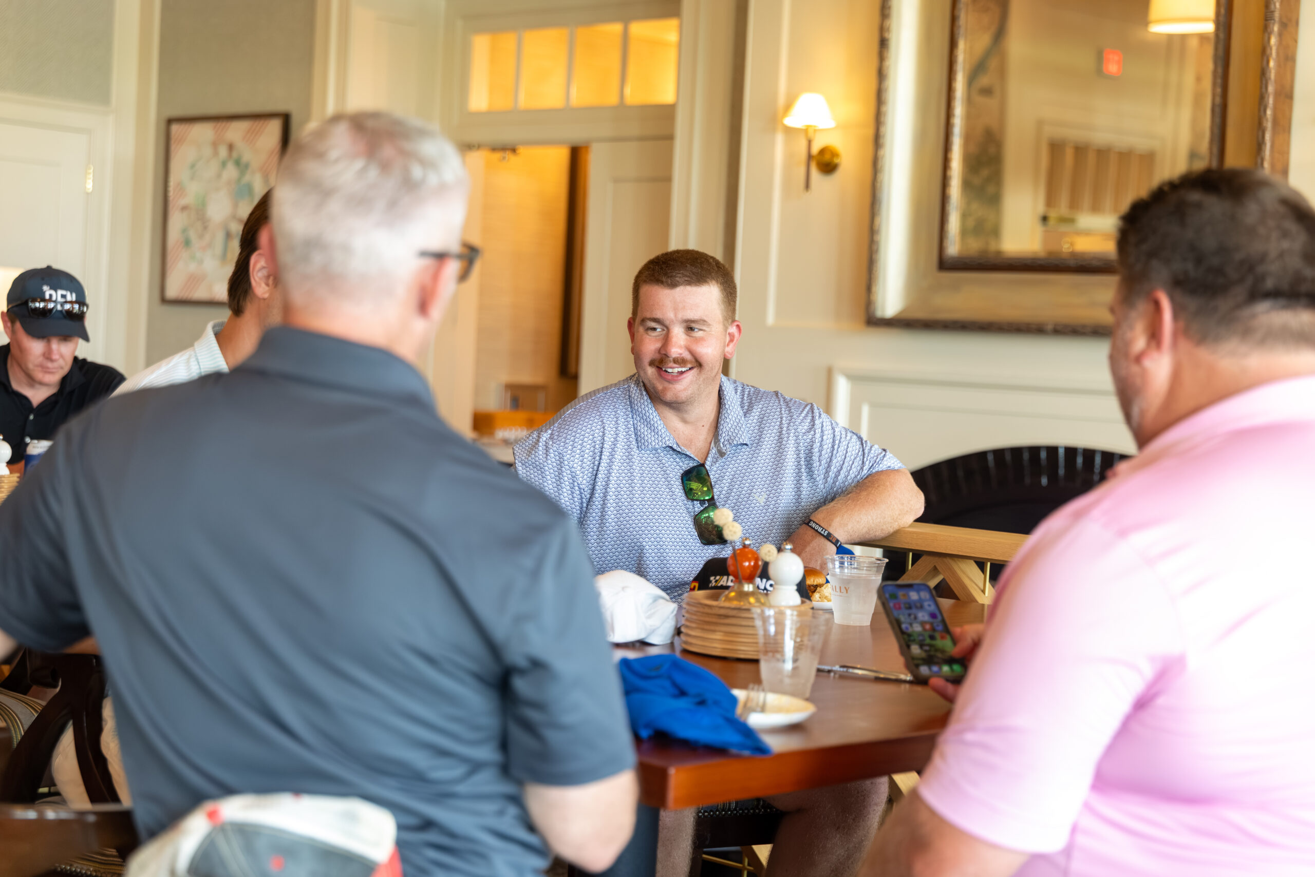 A man in a patterned shirt smiles while talking to two men at a table in a restaurant setting.