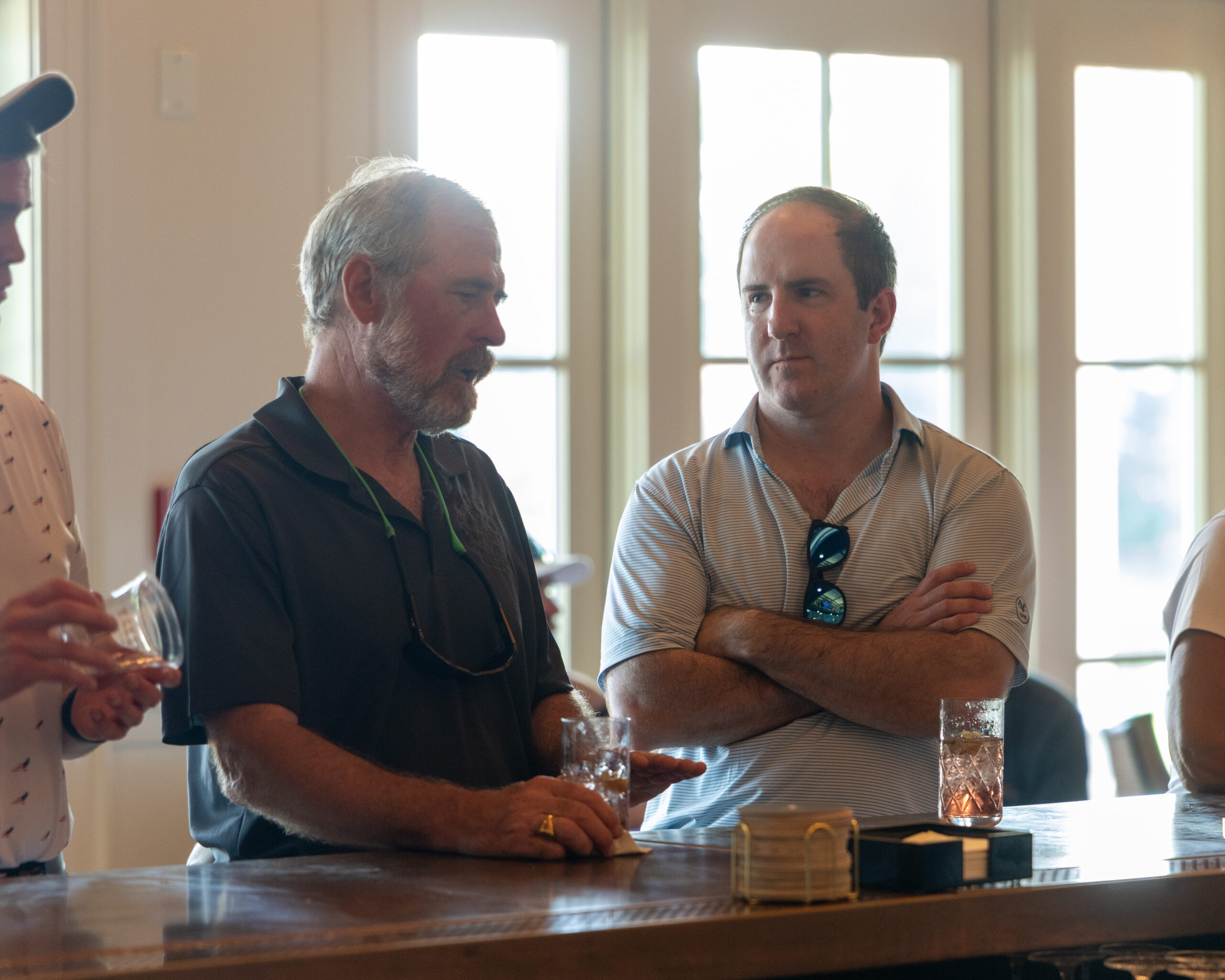 Two men and part of another person stand at a bar, holding drinks.