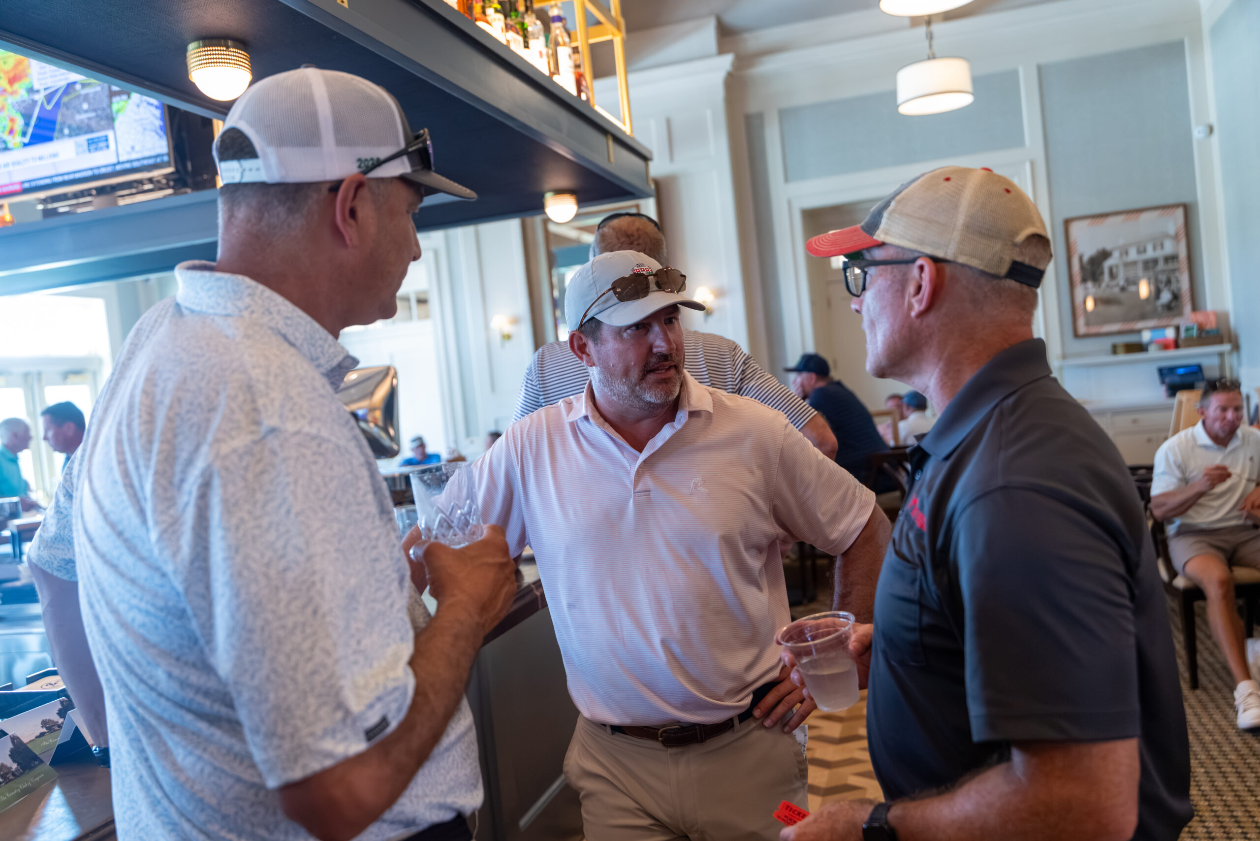 Three men in golf attire with hats and shirts are standing near a bar, talking.