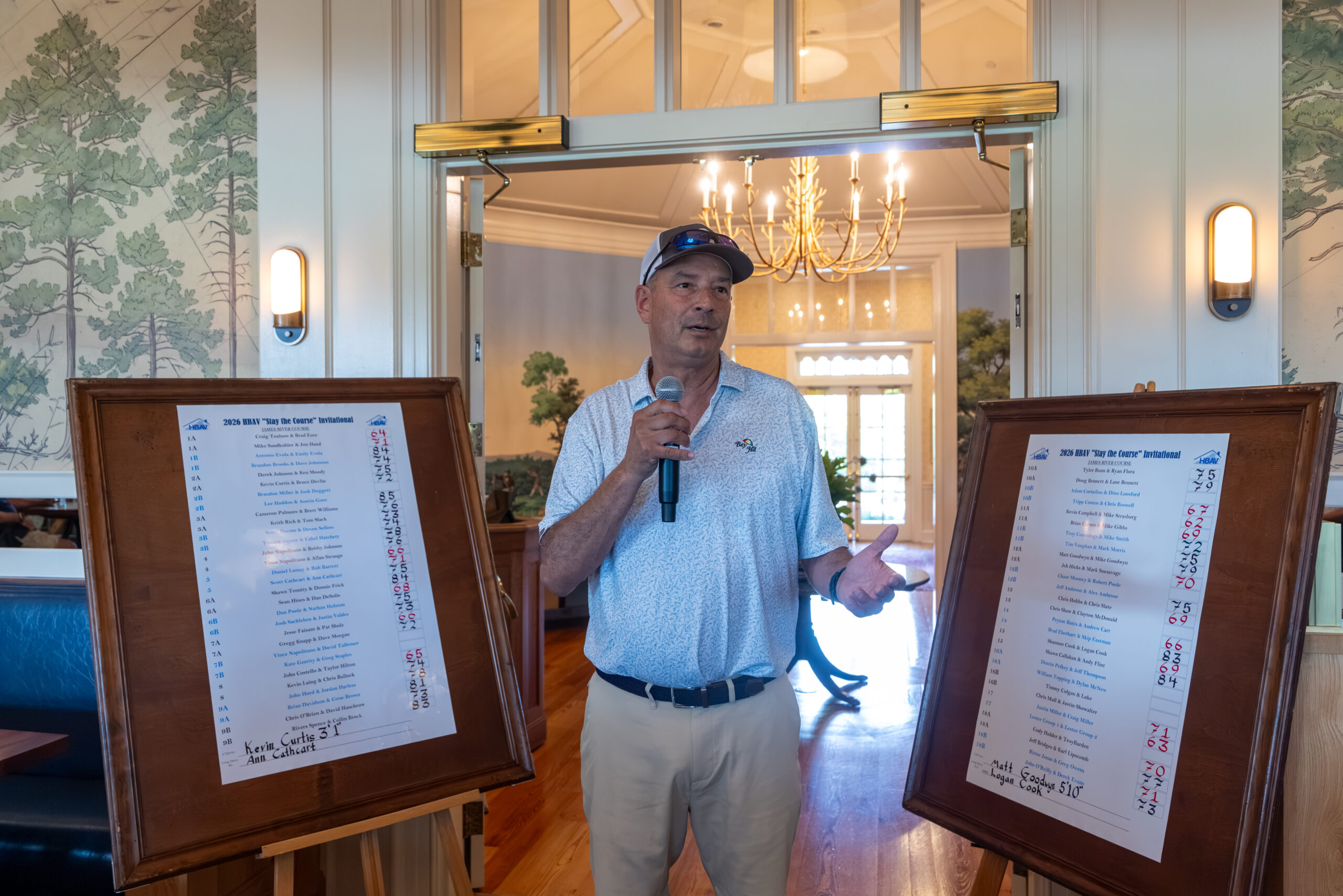 A man holding a microphone and standing between two framed scoreboards displaying a tournament list.