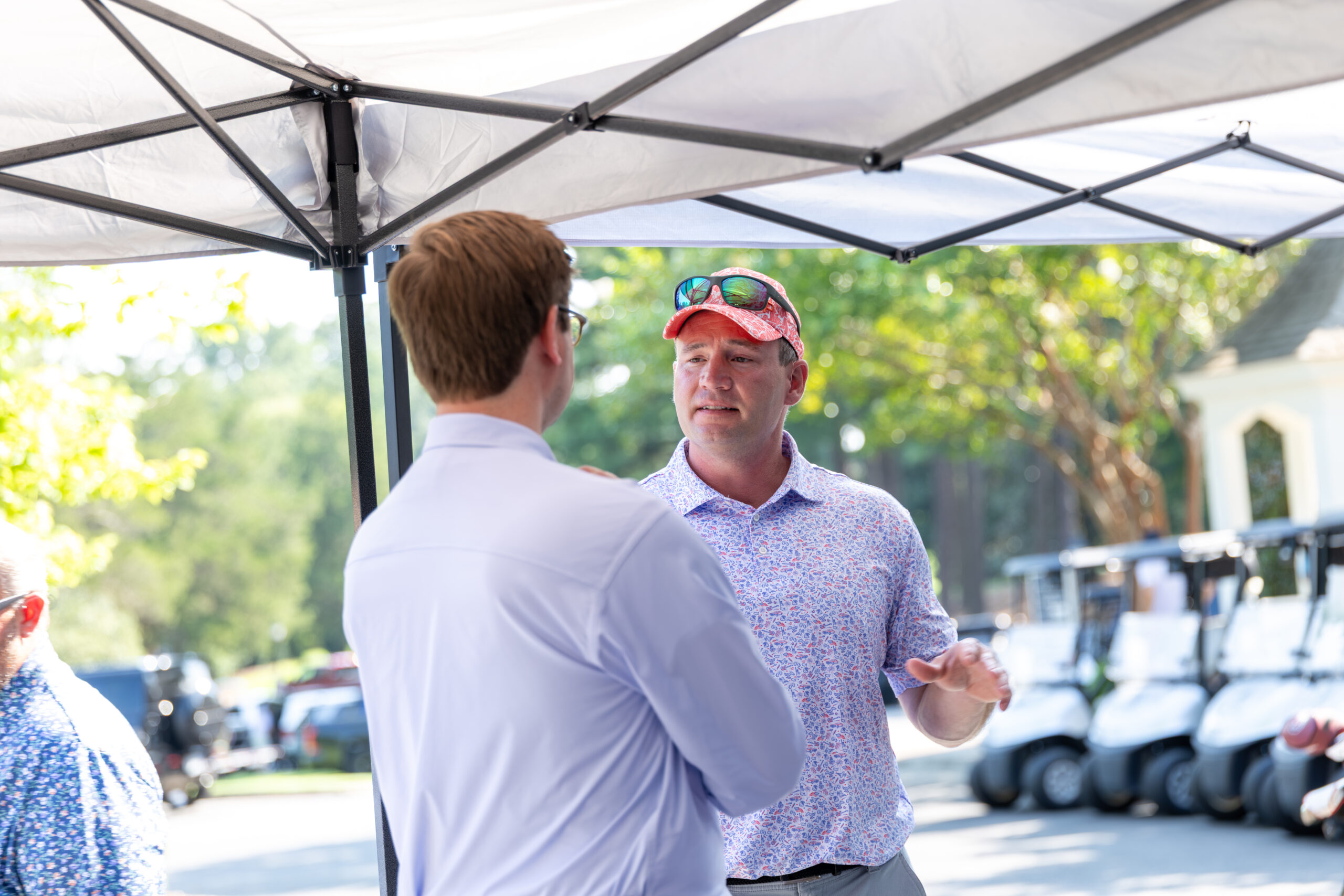 Two men talking under a white canopy tent at a golf course.