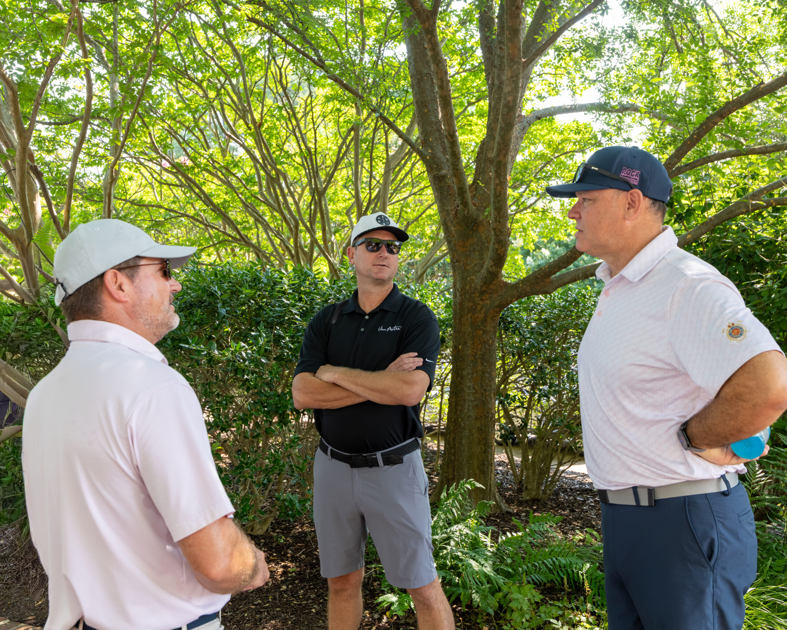 Three men standing and talking outdoors under a large tree with green leaves.