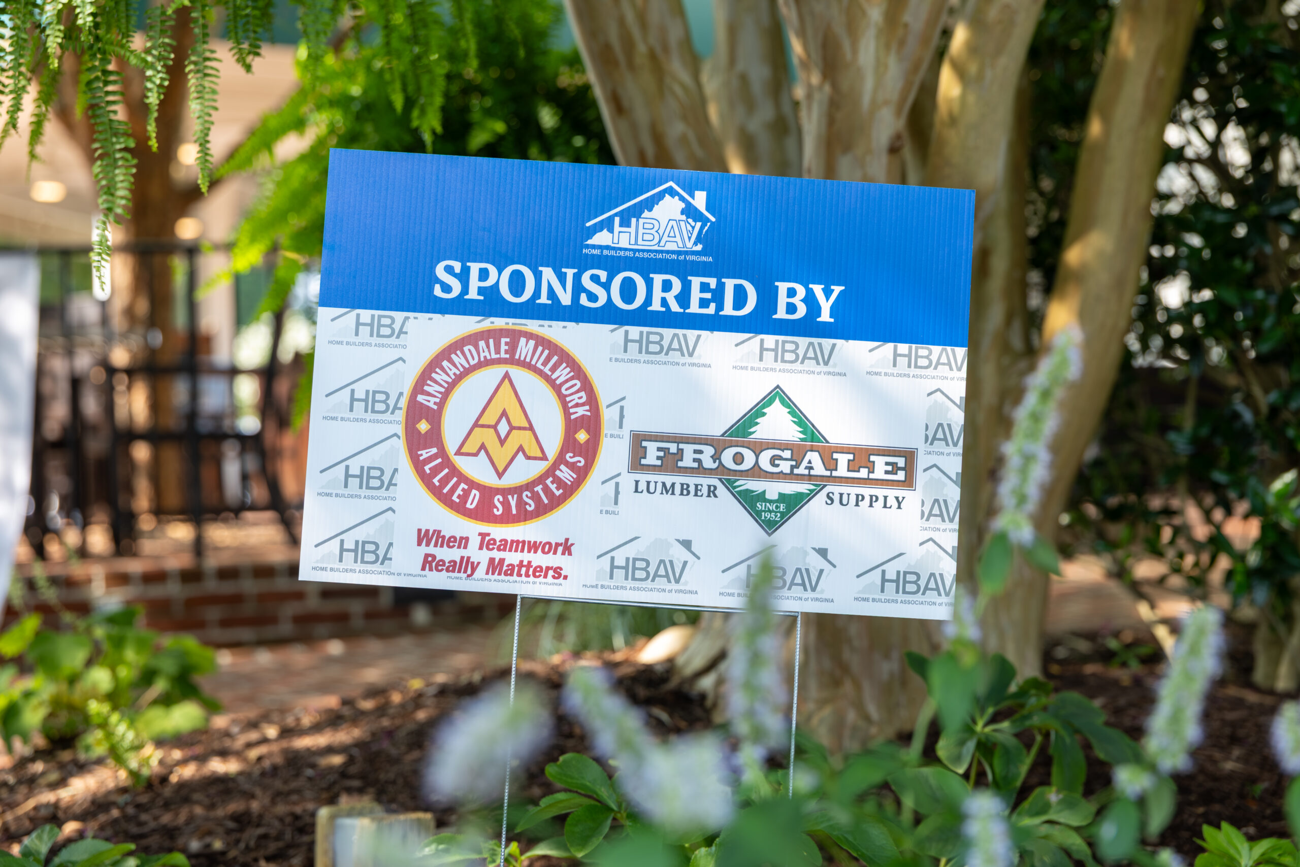 A blue and white sign for HBAV and other sponsors is held by two thin metal poles among garden plants.