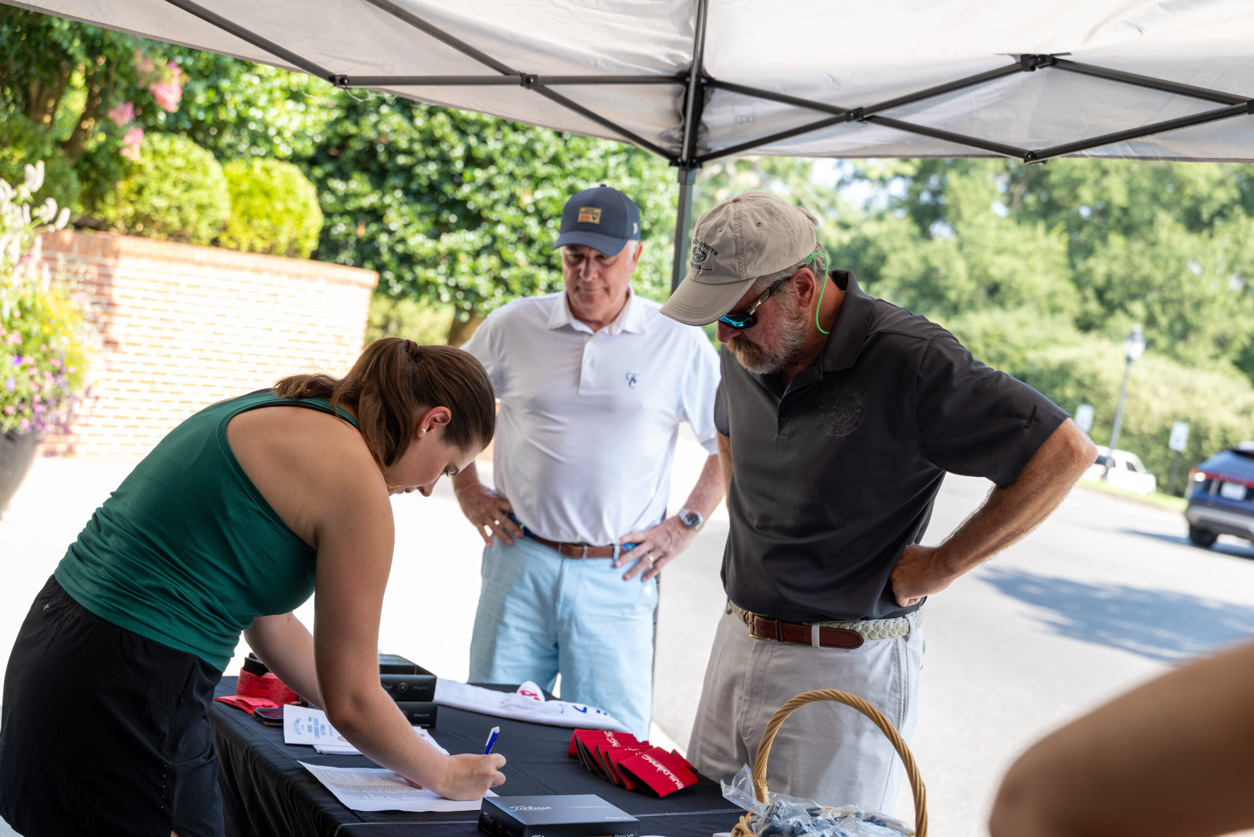 A woman and two men under a white canopy tent at an outdoor event, looking at a table with papers.