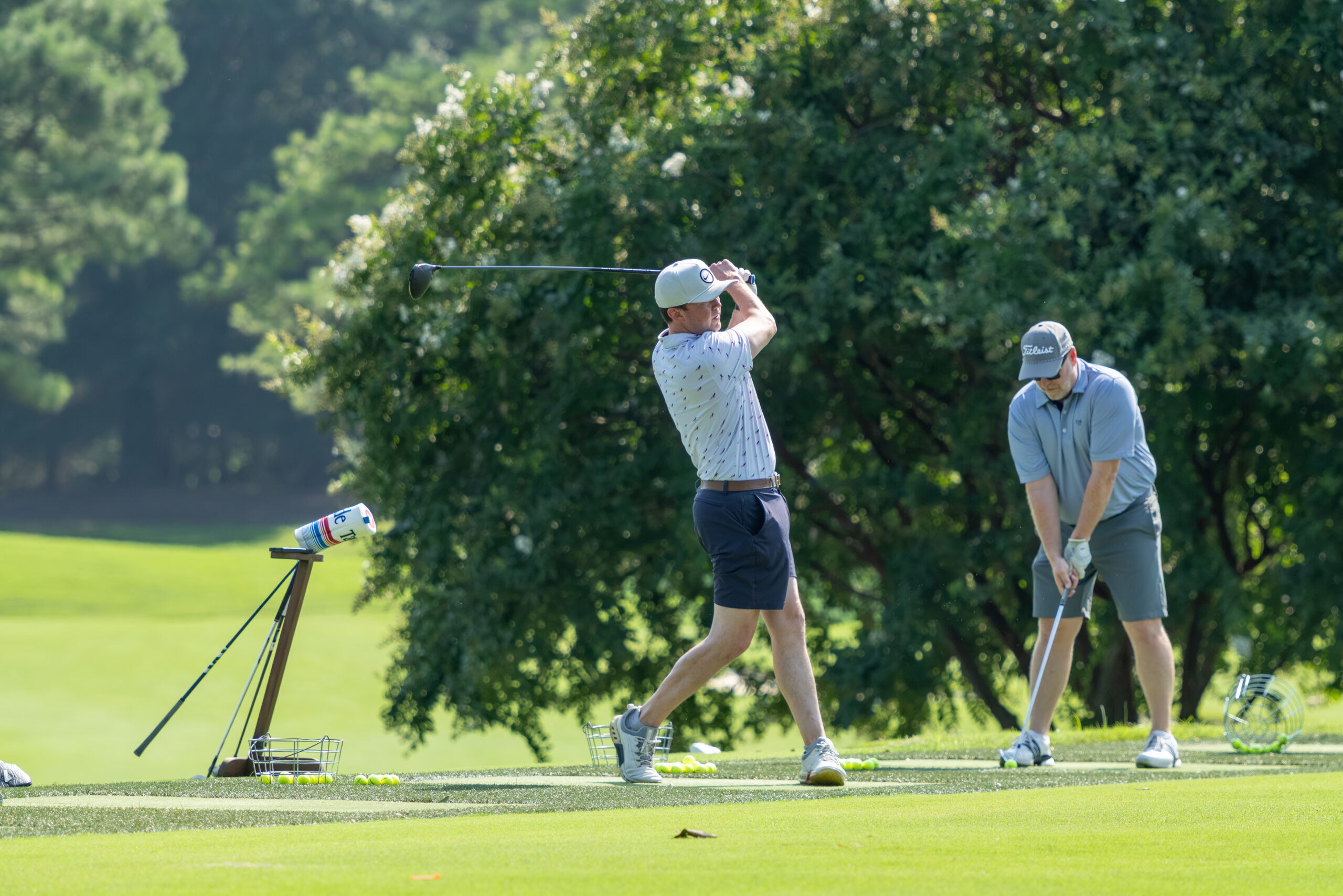 Two men golfing on a grass course with trees in the background.