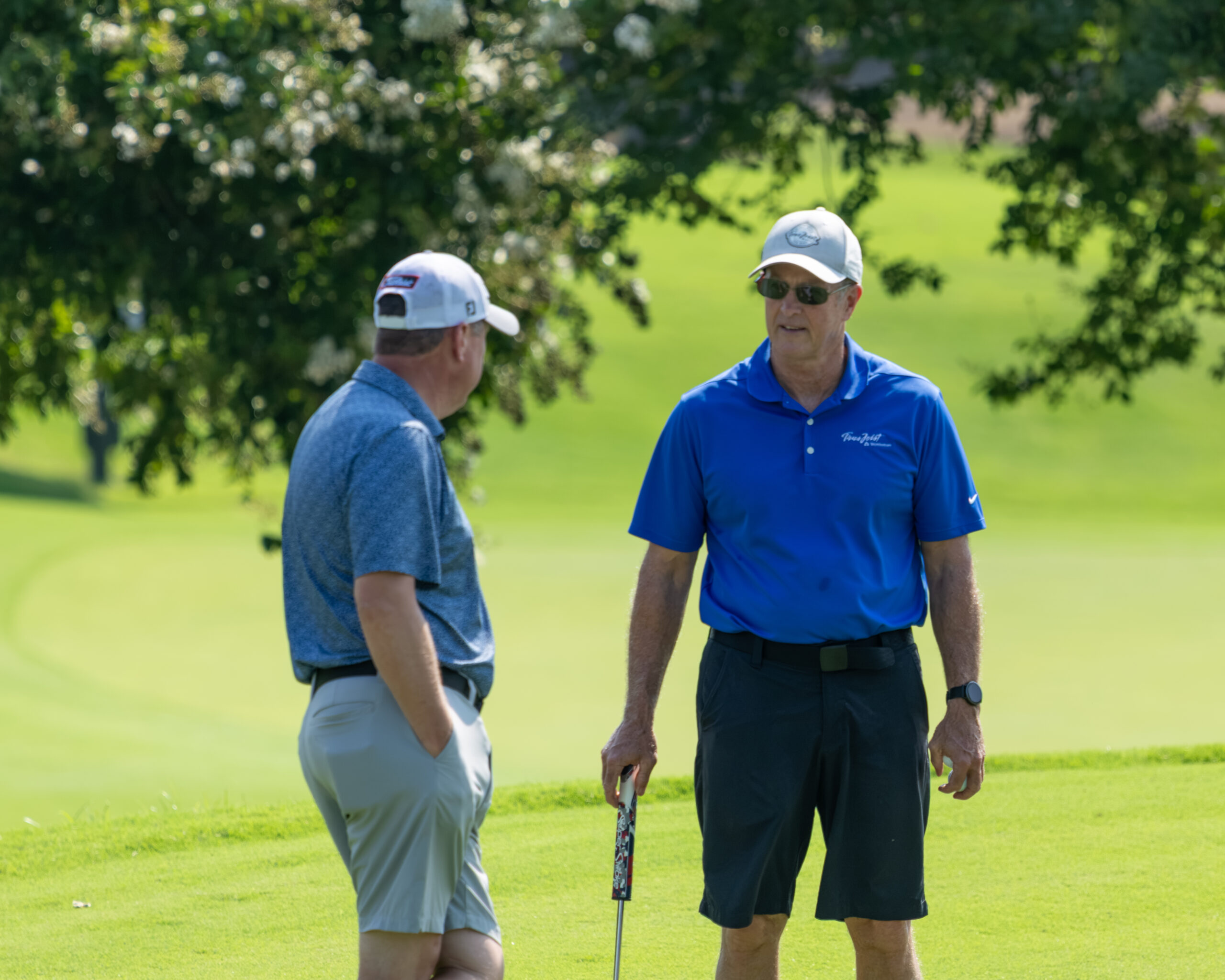 Two men in golf attire standing on a grass field near a tree.