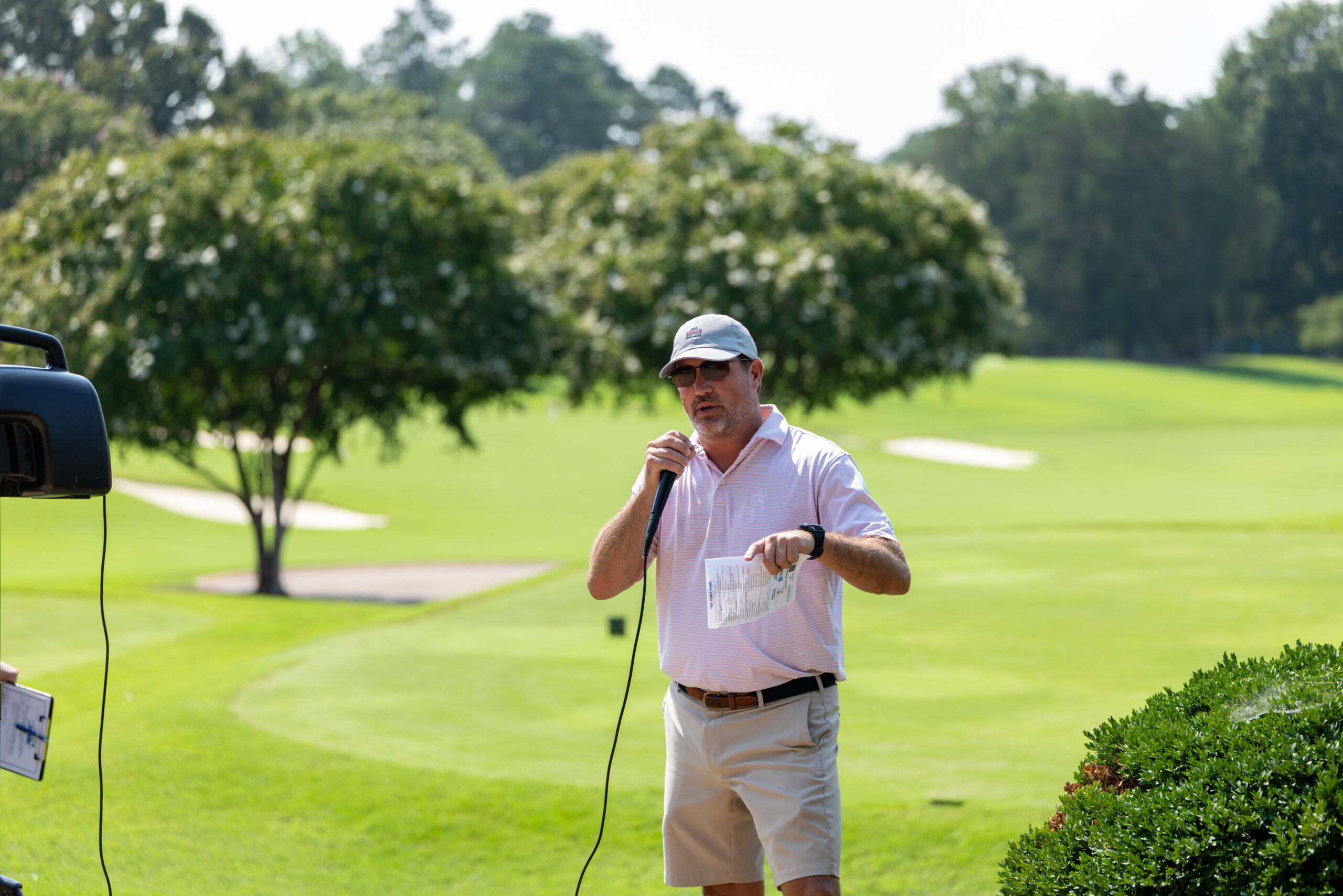 A man wearing a white cap and pink shirt holds a microphone and notes on a golf course.